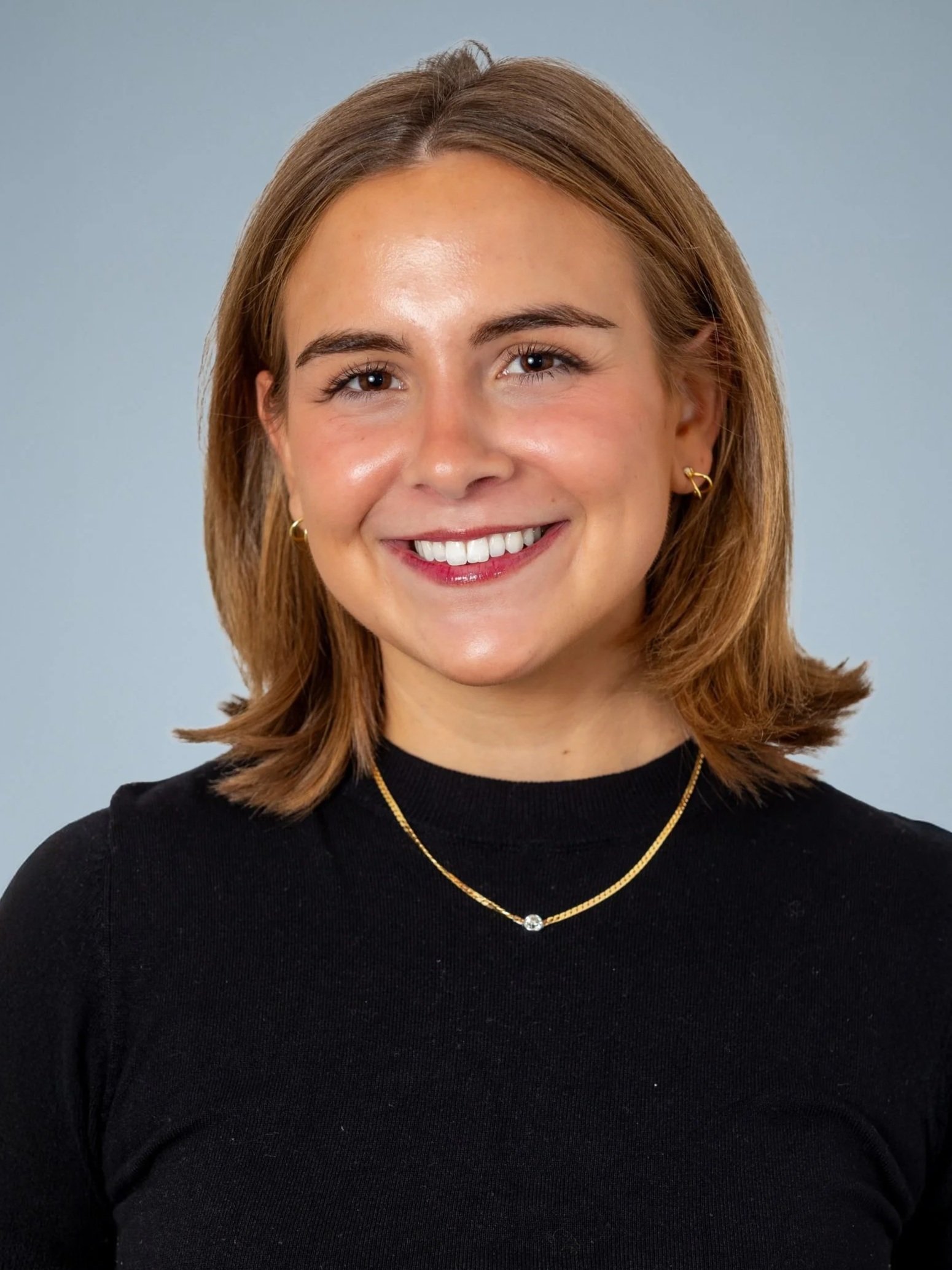 A young woman with shoulder-length light brown hair, smiling, wearing a black top, gold jewelry including small earrings, a necklace with a small gemstone, and a delicate gold chain, against a gray background.