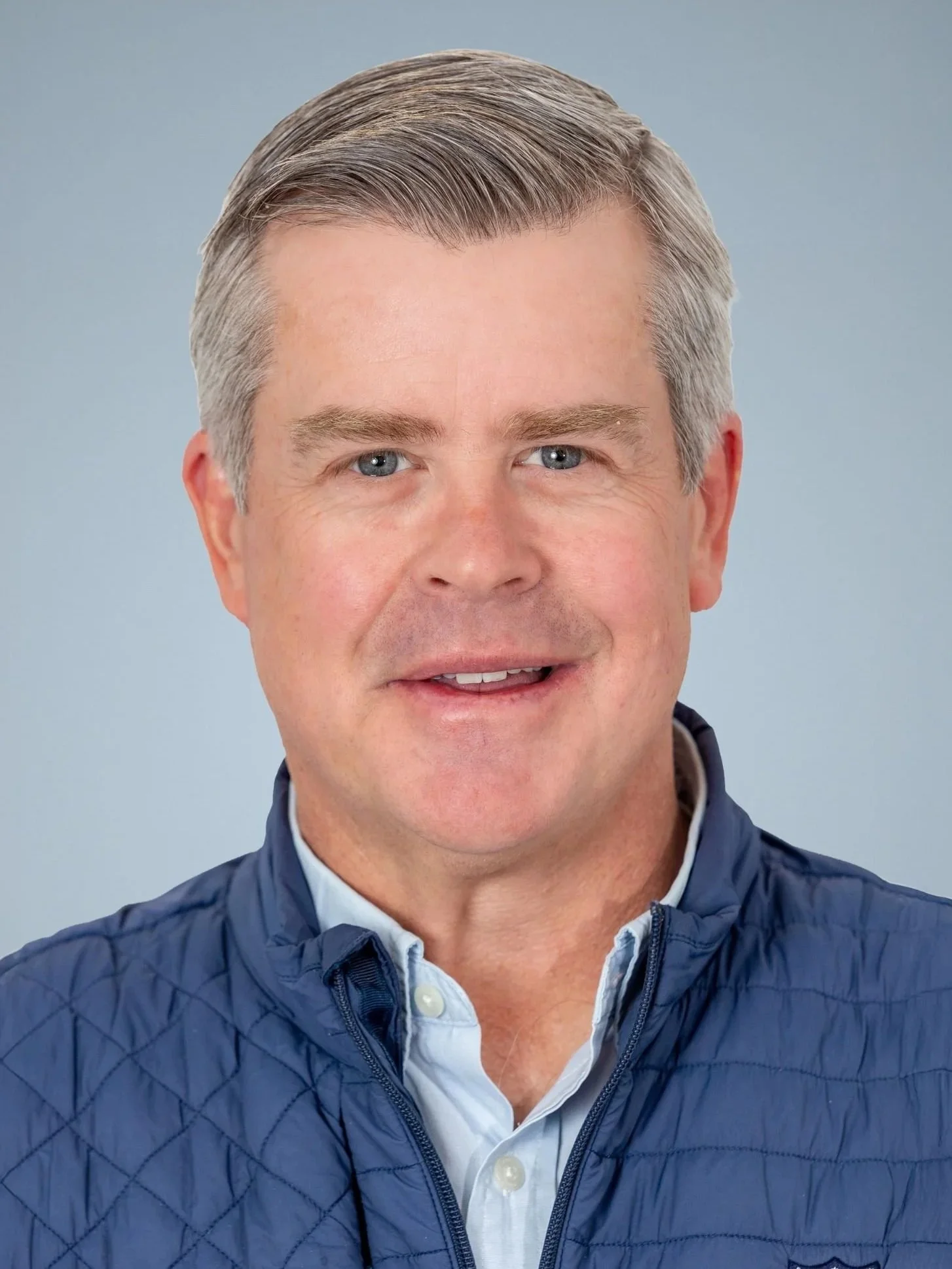 Headshot of a man with gray hair wearing a blue quilted jacket and a light blue button-up shirt, smiling at the camera.