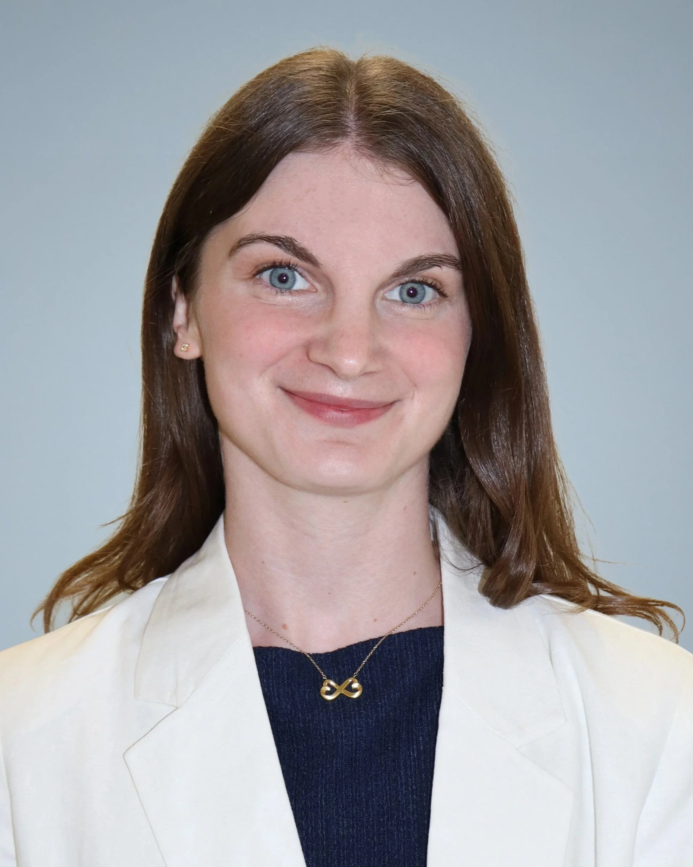 A woman with light skin and blue eyes smiling at the camera, wearing a white blazer, a dark top, and a gold infinity necklace.