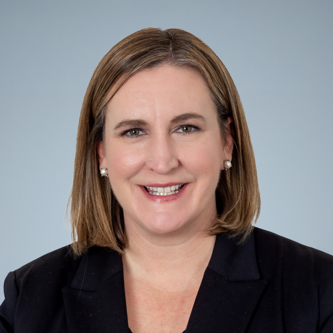 Headshot of a woman with shoulder-length brown hair, wearing a black blazer, pearl earrings, and smiling against a light blue background.