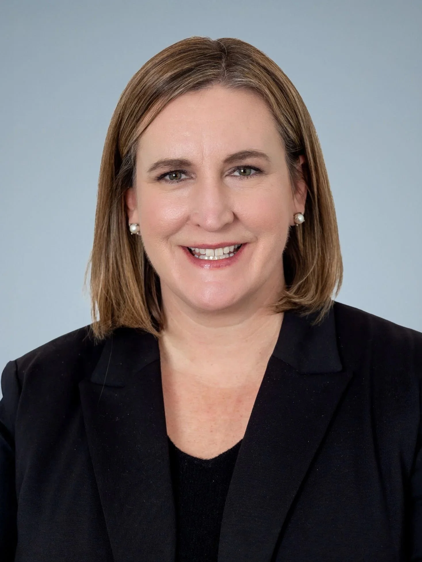 A woman with shoulder-length brown hair, wearing a black blazer and pearl earrings, smiling against a plain gray background.
