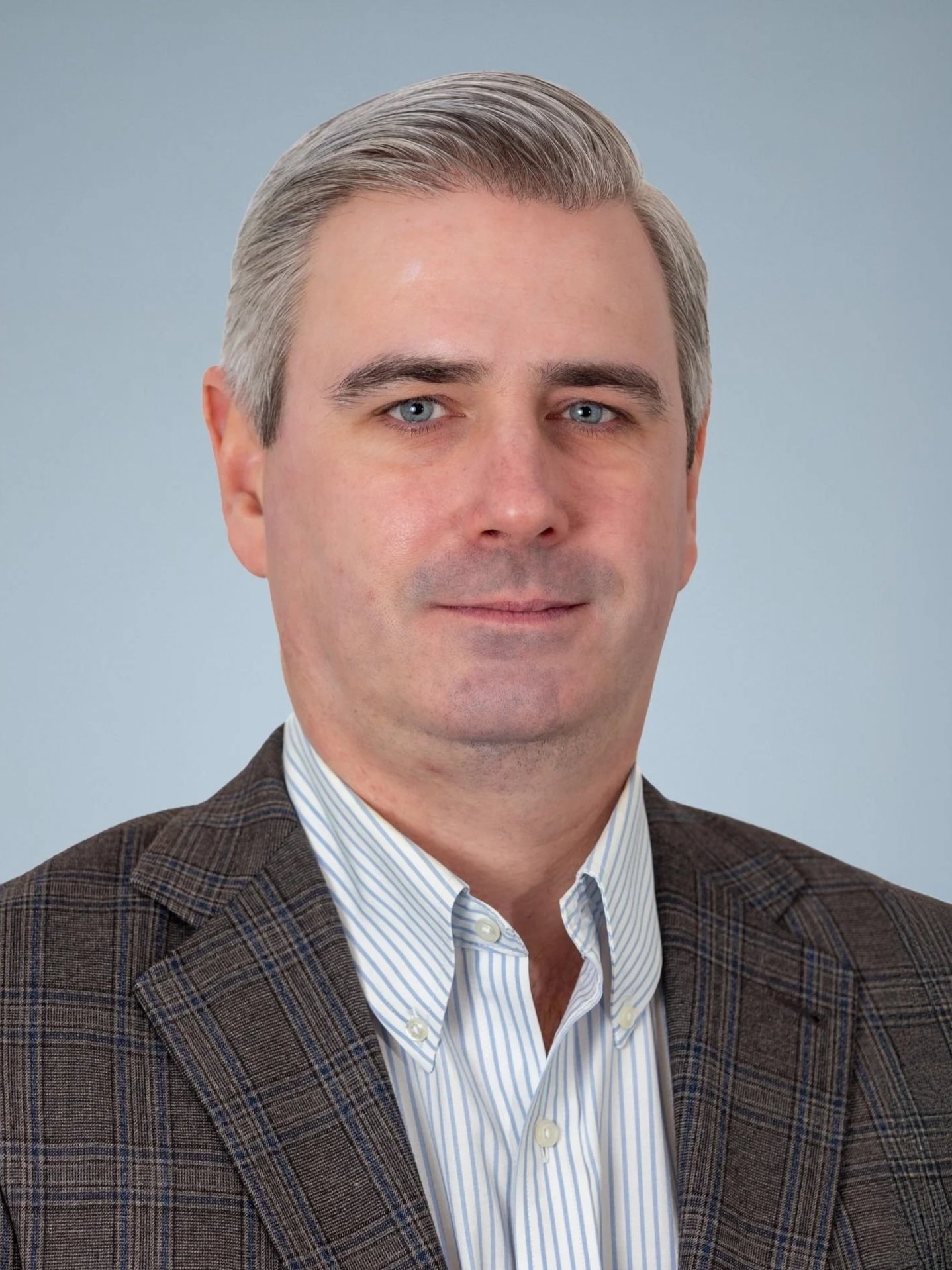 A professional headshot of a man with short gray hair, blue eyes, wearing a checkered brown blazer and a light striped dress shirt, against a plain light blue background.