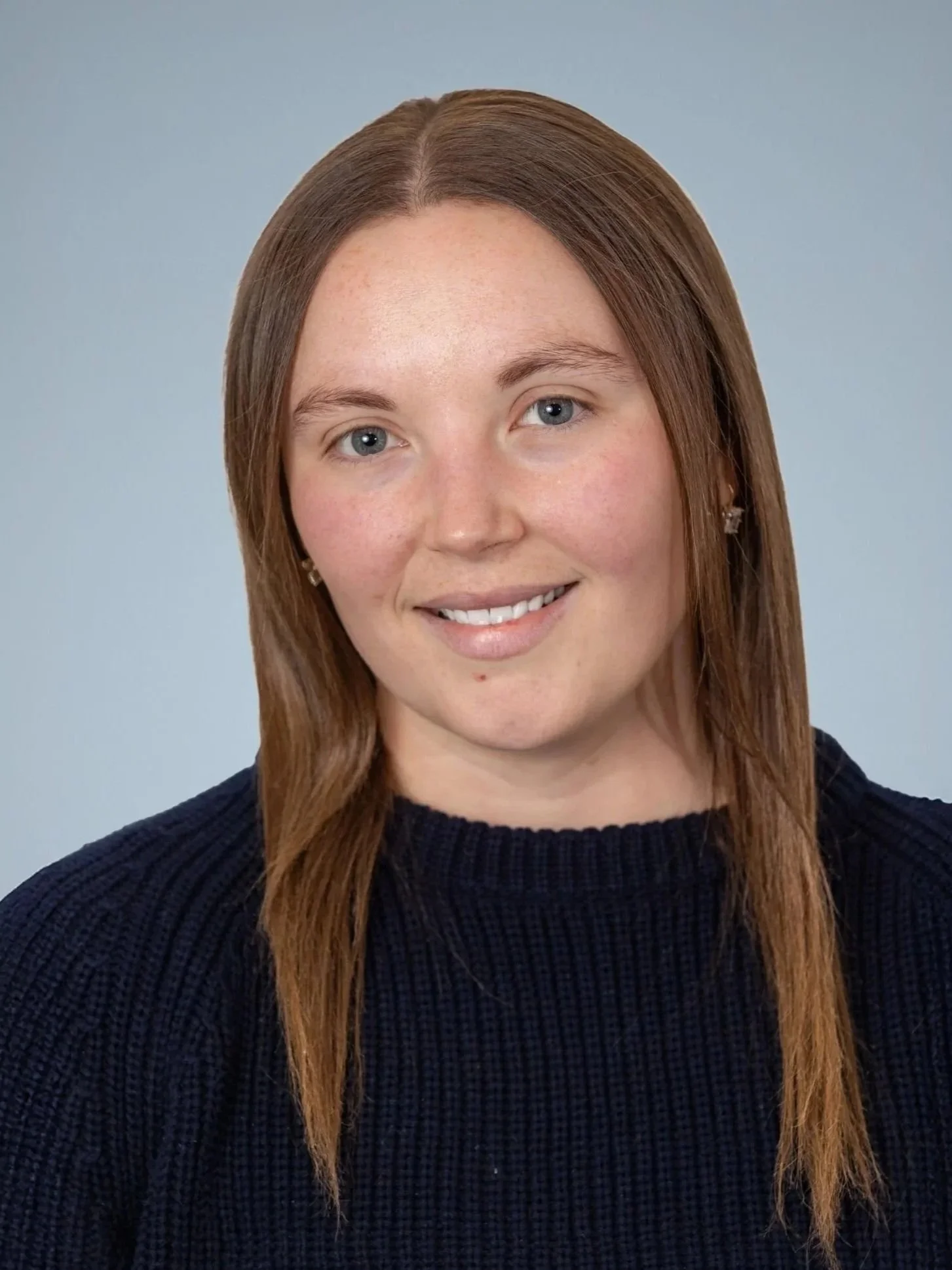 Headshot of a woman with straight, shoulder-length reddish-brown hair, blue eyes, and light skin, smiling, wearing a dark knit top and small star-shaped earrings, against a neutral gray background.