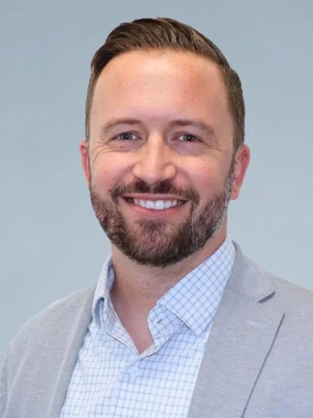 Portrait of a smiling man with a beard, wearing a light gray blazer and white checkered shirt against a plain light background.