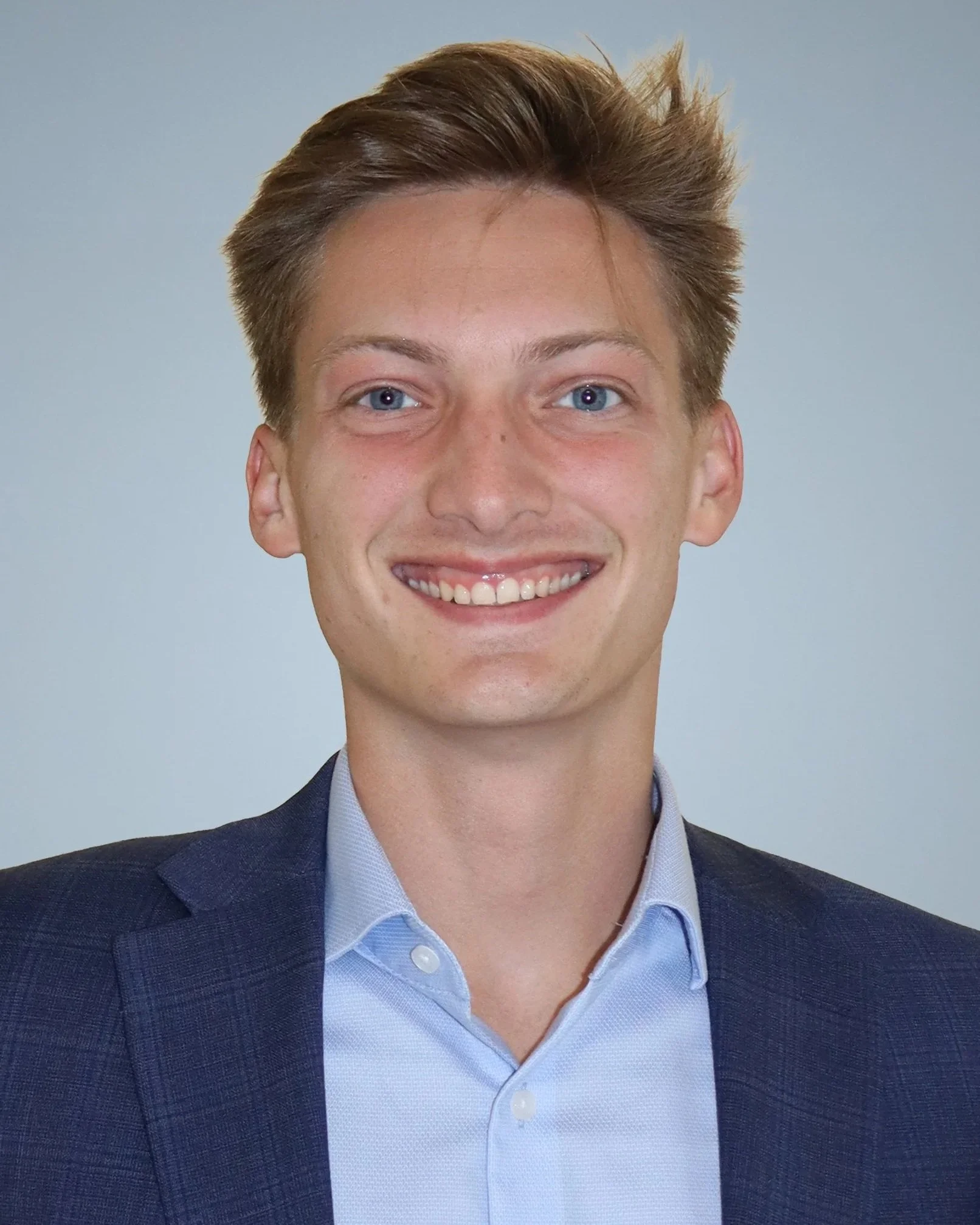 A young man with short, light brown hair, blue eyes, and a wide smile, wearing a dark suit jacket and light blue dress shirt, posing against a plain light grey background.