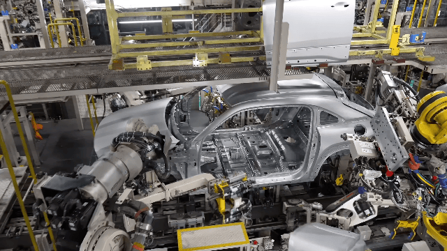 Automobile assembly line with a partially assembled silver car body and robotic arms assembling parts.