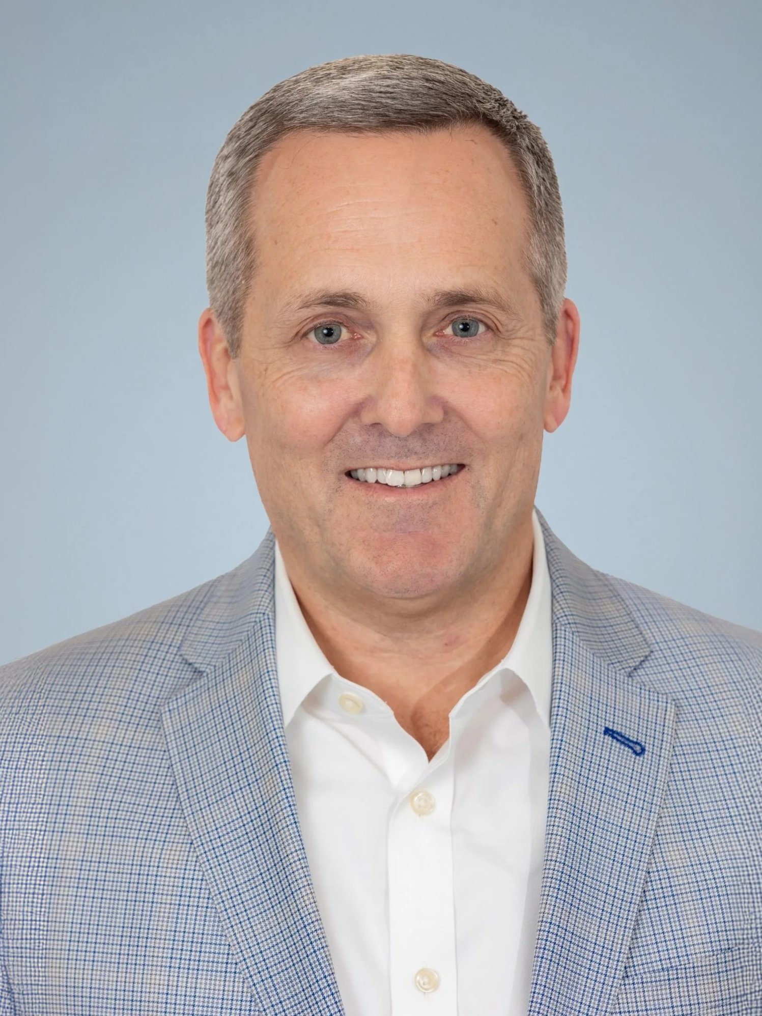 Close-up of a smiling middle-aged man with gray hair, wearing a light-colored blazer and white dress shirt against a light gray background.
