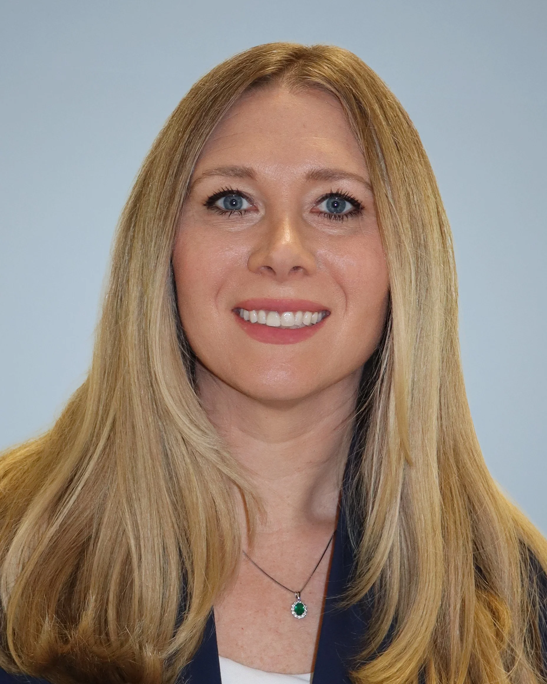 A woman with long blonde hair, blue eyes, and fair skin smiling, wearing a black top with a necklace featuring a green gemstone pendant, against a light blue background.