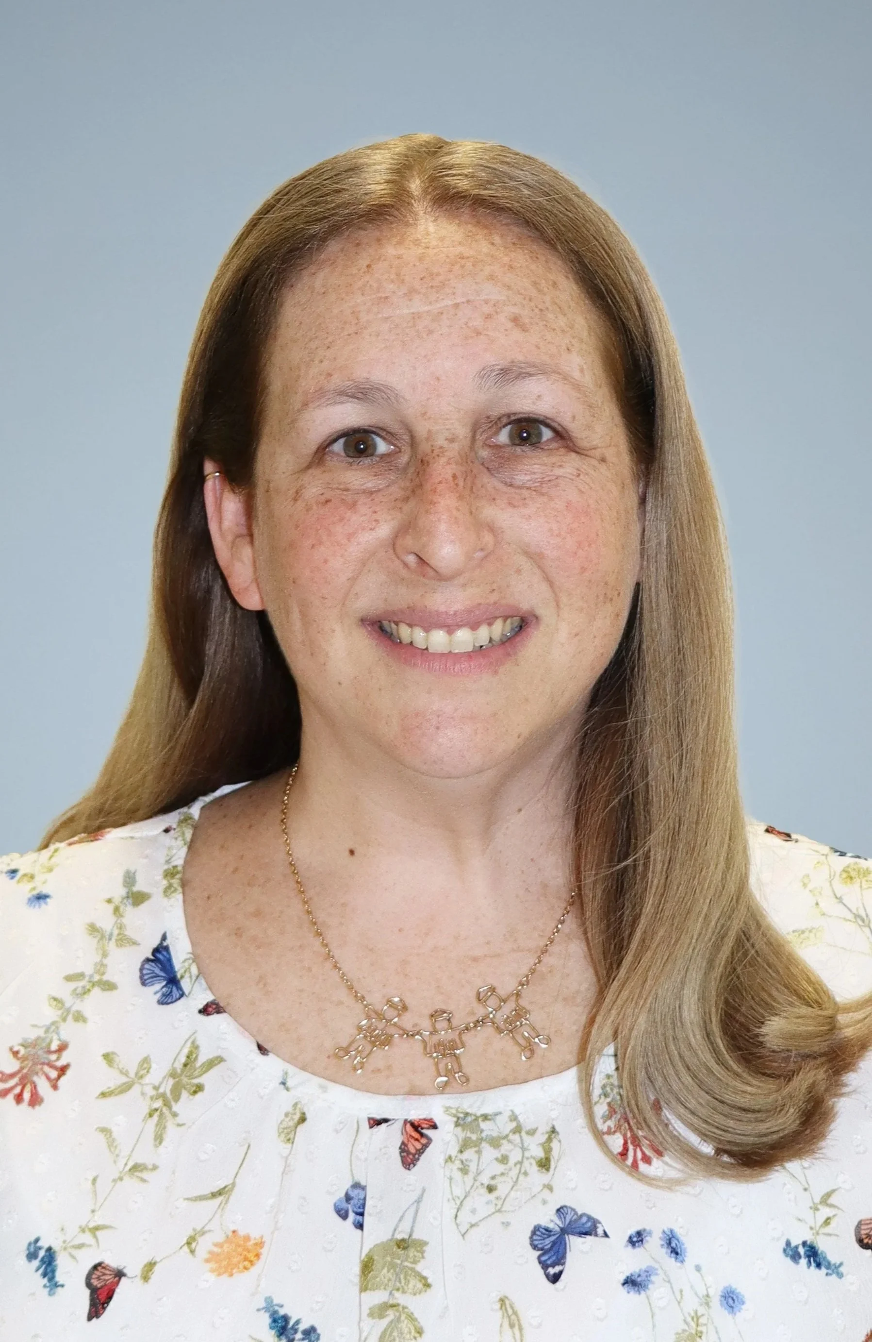 A woman with long red hair, freckles, and a light complexion smiling at the camera, wearing a patterned blouse and a gold necklace with a name pendant.