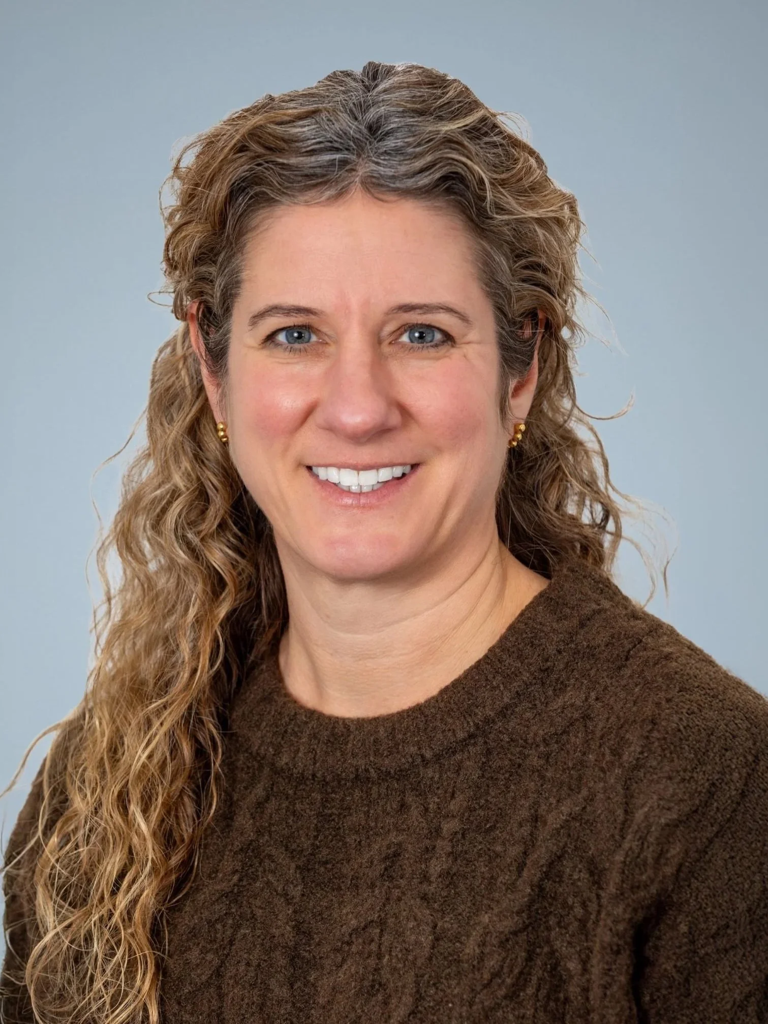 Portrait of a woman with long curly hair and blue eyes, wearing a brown sweater, smiling against a light blue background.