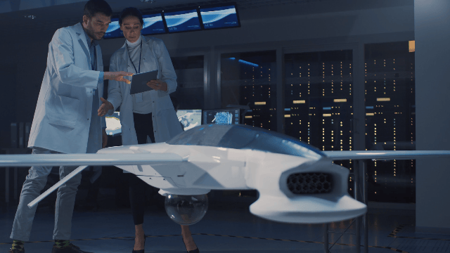 Two scientists in lab coats examining a large drone or unmanned aircraft in a high-tech facility with multiple monitors and server racks.