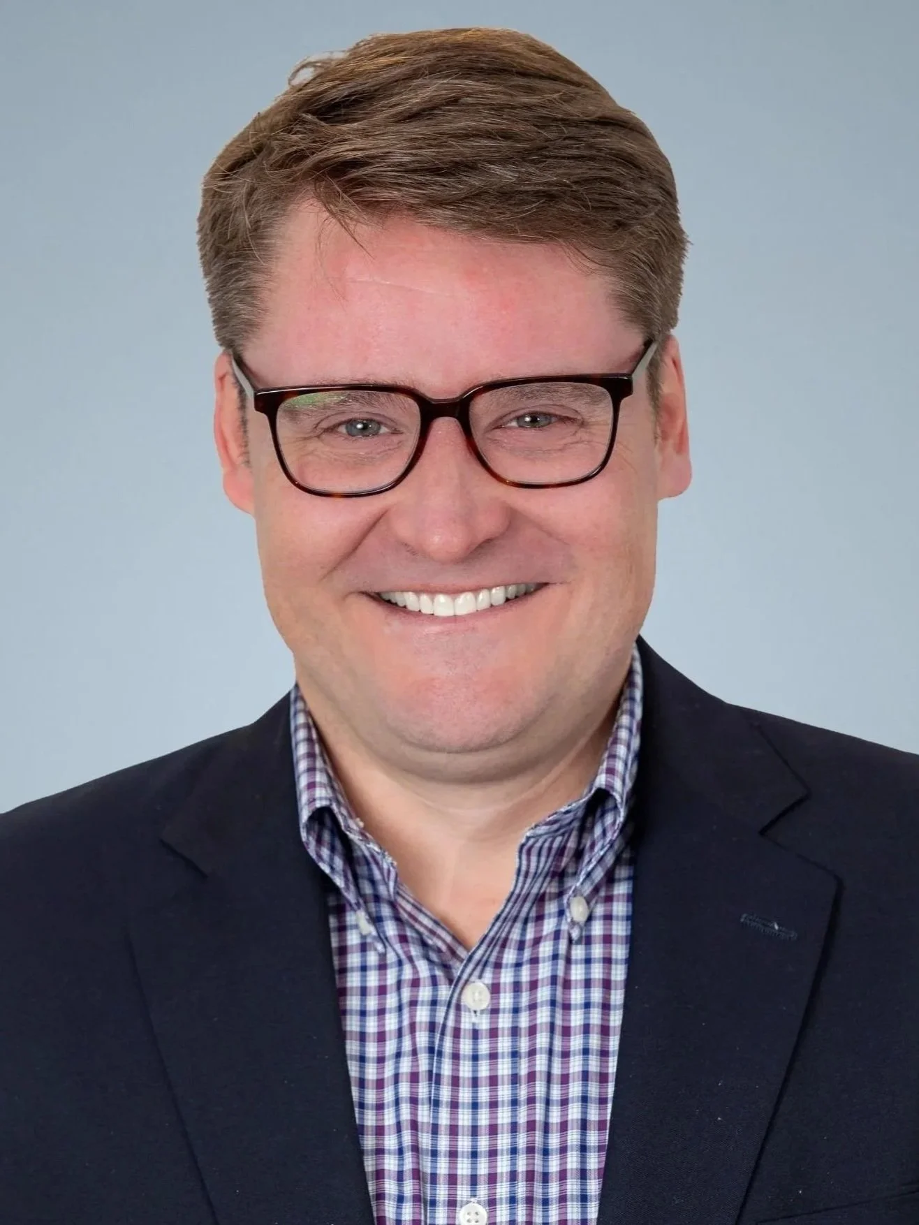 Headshot of a smiling man with light brown hair, glasses, wearing a dark blazer and a checkered shirt against a plain background.