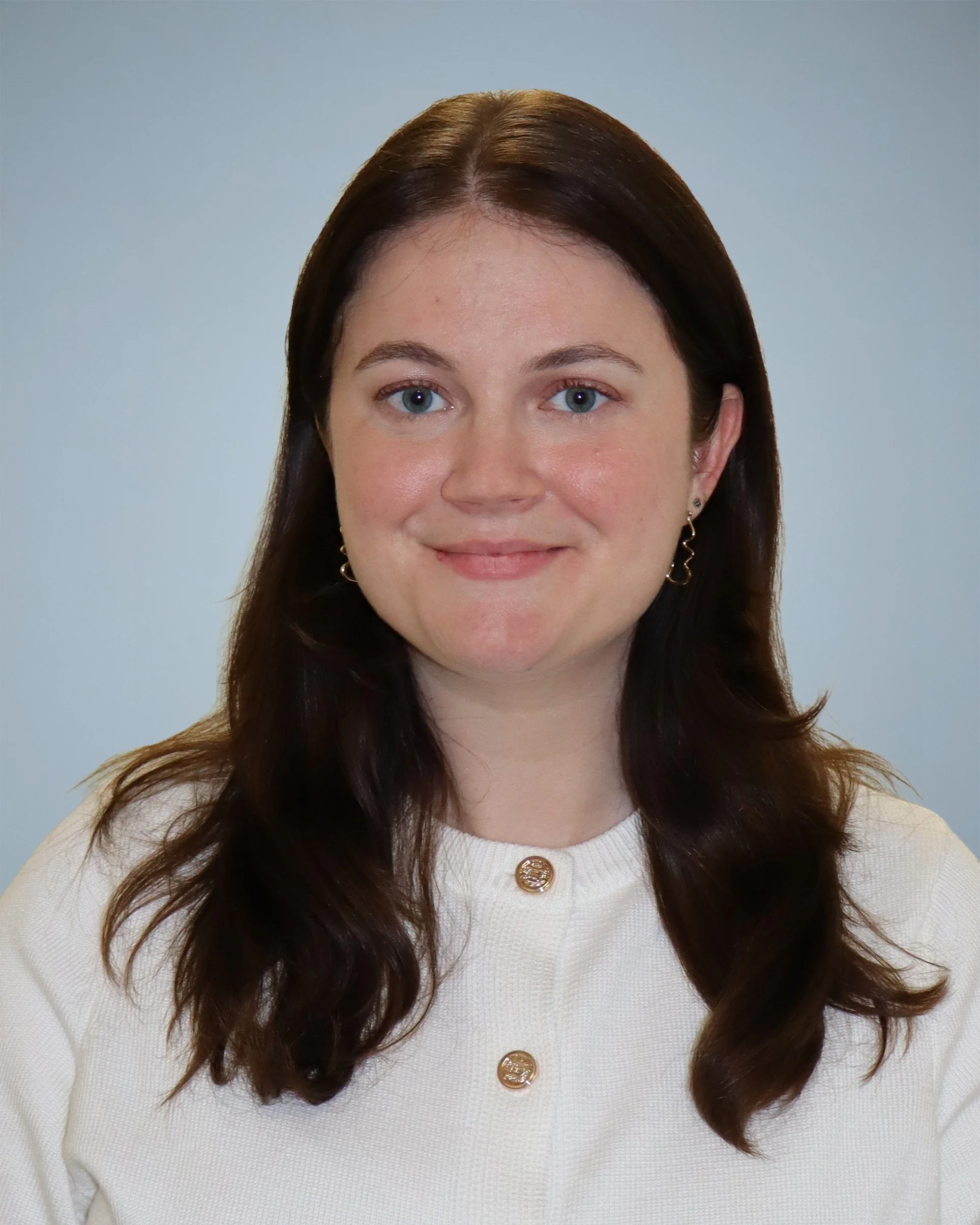 Close-up of a woman with long, wavy dark hair wearing a white top with gold buttons, gold earrings, and smiling against a light gray background.