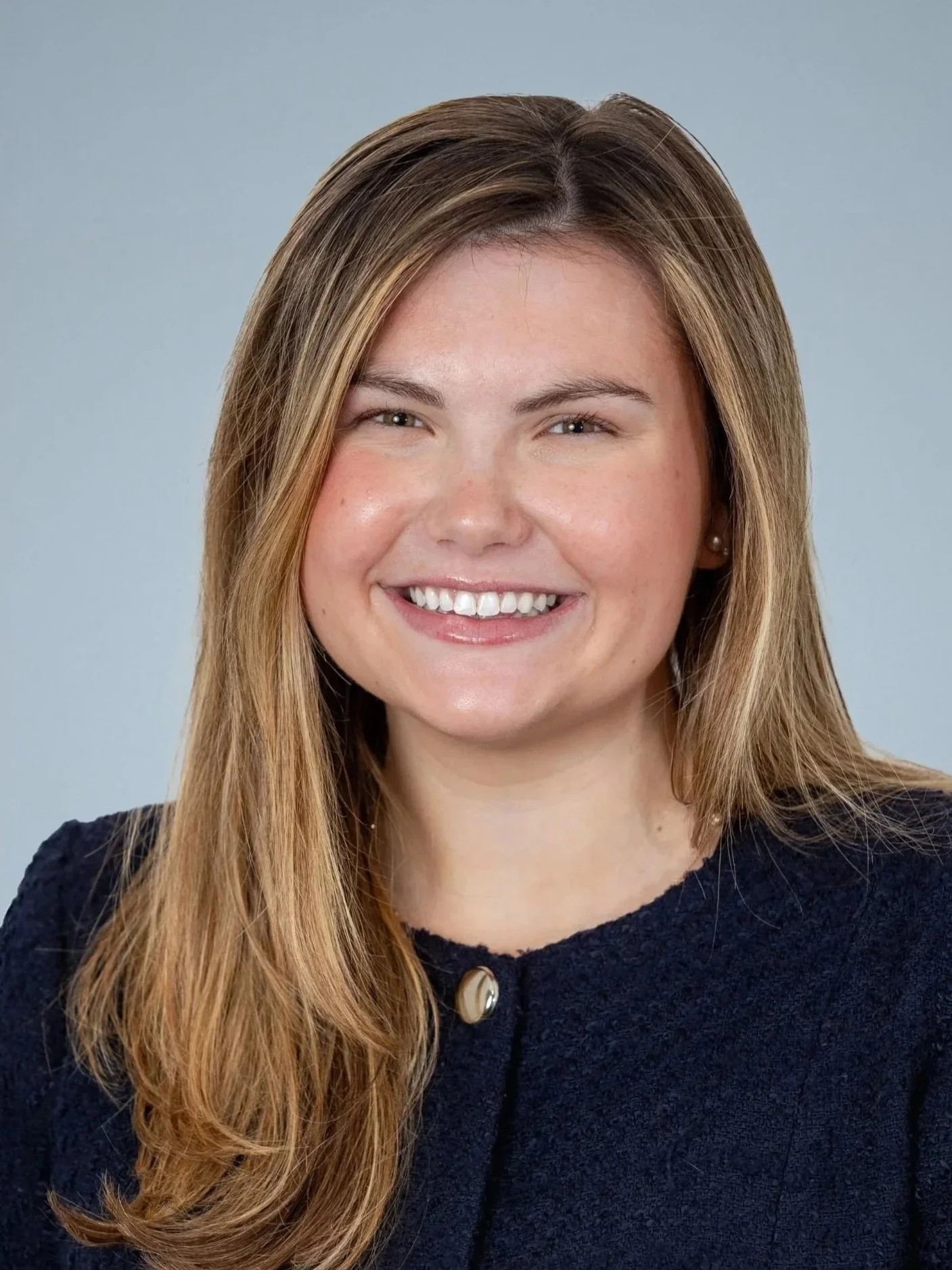 Close-up of a young woman with long, light brown hair, smiling, wearing a dark sweater with a pearl button, and small pearl earrings, against a light gray background.
