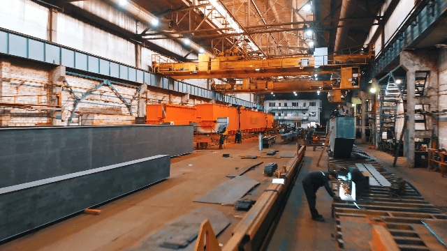 Interior of a large industrial warehouse with workers welding large metal structures and a yellow overhead crane.