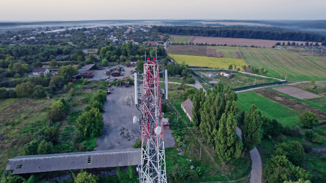 A tall communication tower with multiple antennas stands in a rural area with trees, fields, and buildings. Forest and water body visible in the background.