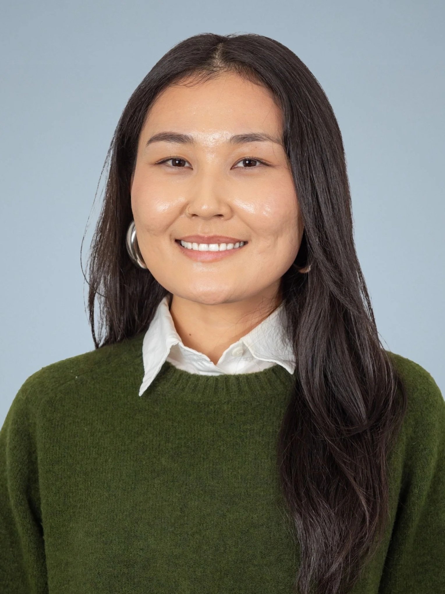 Close-up of a woman with long dark hair, wearing a white collared shirt and a green sweater, smiling in front of a light blue background.