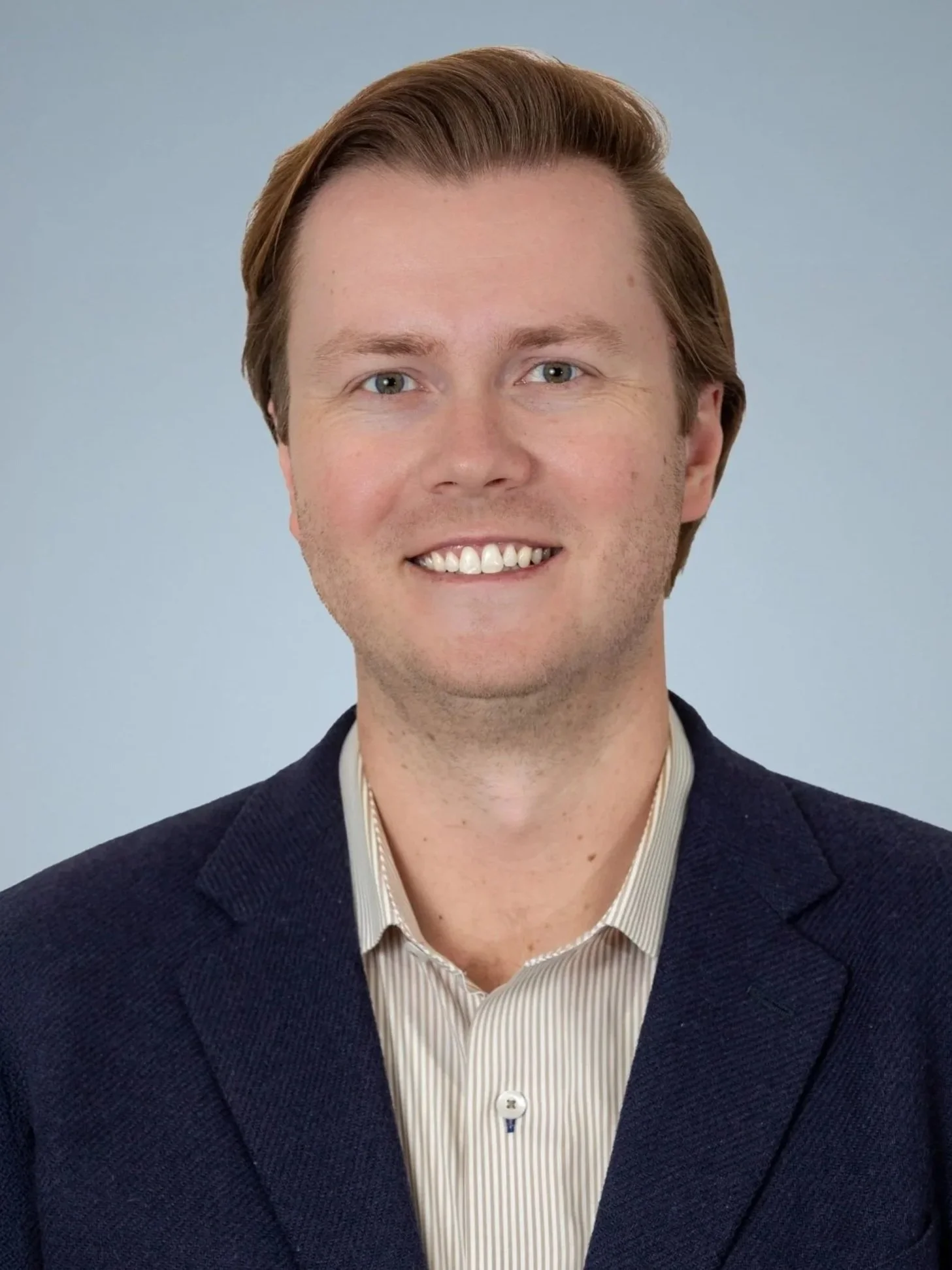 Professional headshot of a young man with light brown hair, blue eyes, and fair skin, wearing a dark blazer and light striped shirt, smiling against a plain background.