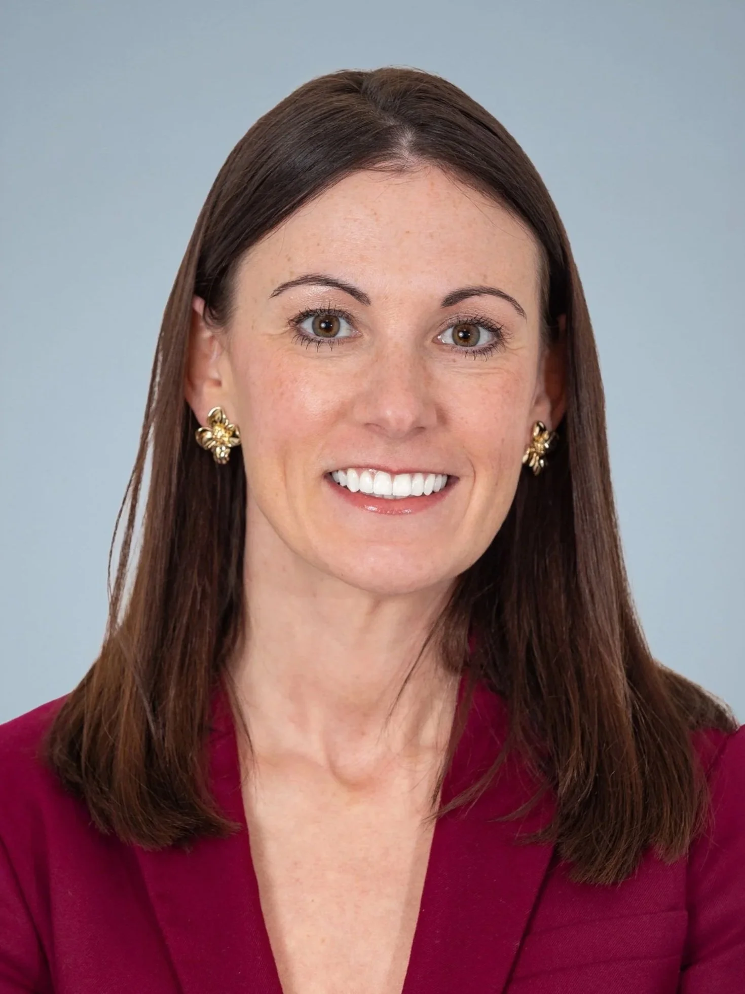 Headshot of a woman with brown hair, wearing gold earrings and a burgundy blazer, smiling against a light grey background.