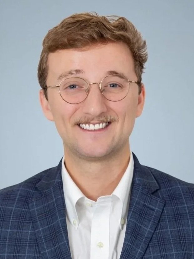 Portrait of a smiling man with glasses, wearing a checkered blazer and white shirt against a plain background.