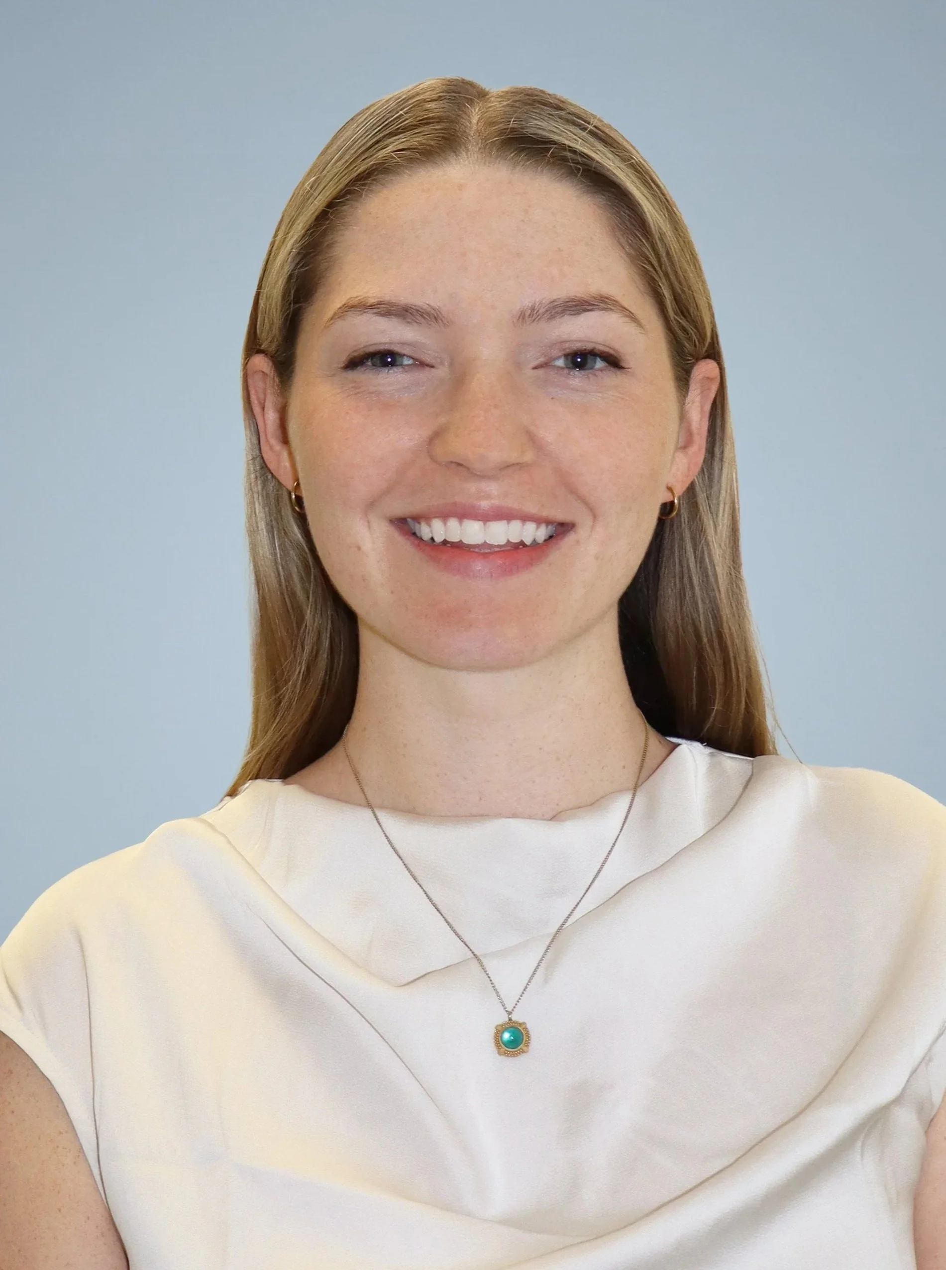 Close-up of a woman with blonde hair wearing a cream-colored top, a necklace with a teal pendant, and small hoop earrings, smiling against a plain background.