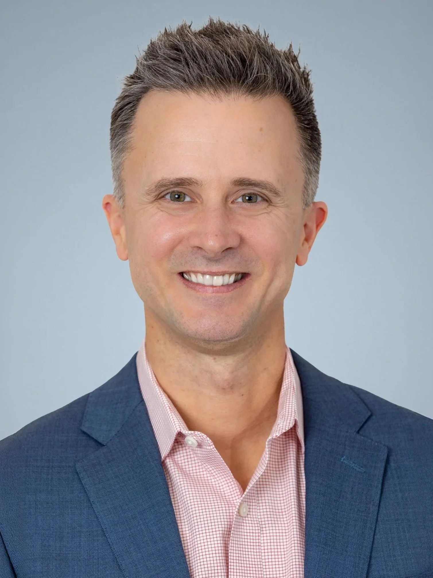 A smiling man in a dark blue blazer and pink checkered shirt against a light blue background.