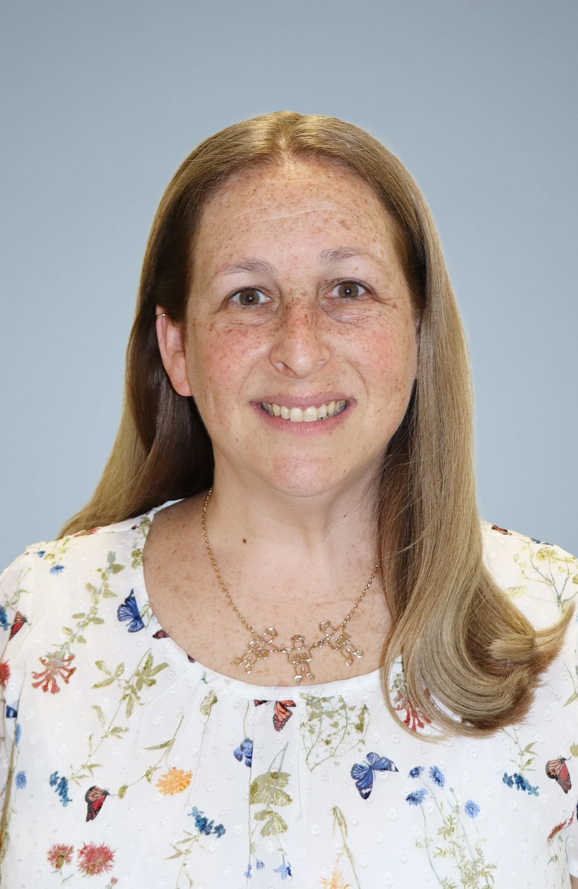 A woman with long, light brown hair and freckles, smiling, wearing a white floral dress and a gold necklace, against a plain light blue background.