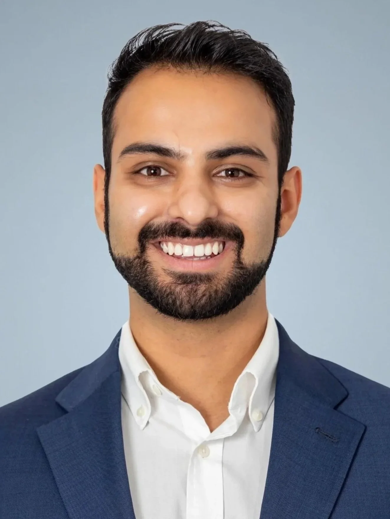 Headshot of a smiling man with dark hair, beard, wearing a navy blue suit and white shirt against a light blue background.