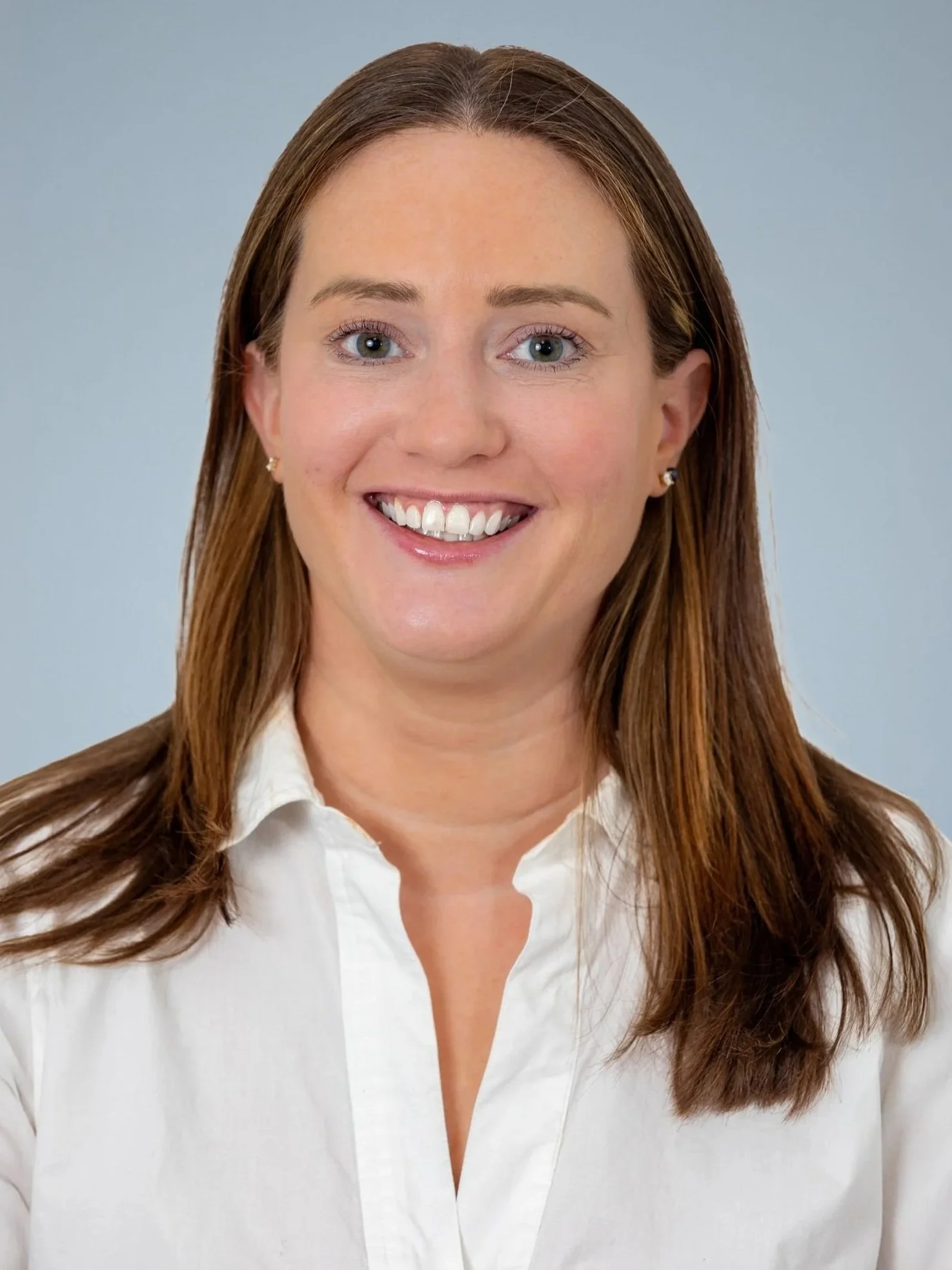 Smiling woman with shoulder-length brown hair wearing a white button-up shirt against a light blue background.