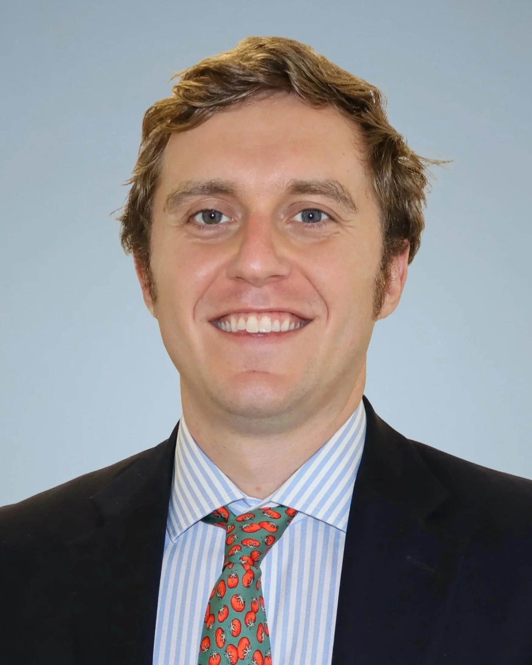 Close-up of smiling young man with light skin, red hair, and wearing a dark suit, light blue striped shirt, and a tie featuring tomatoes, against a plain gray background.