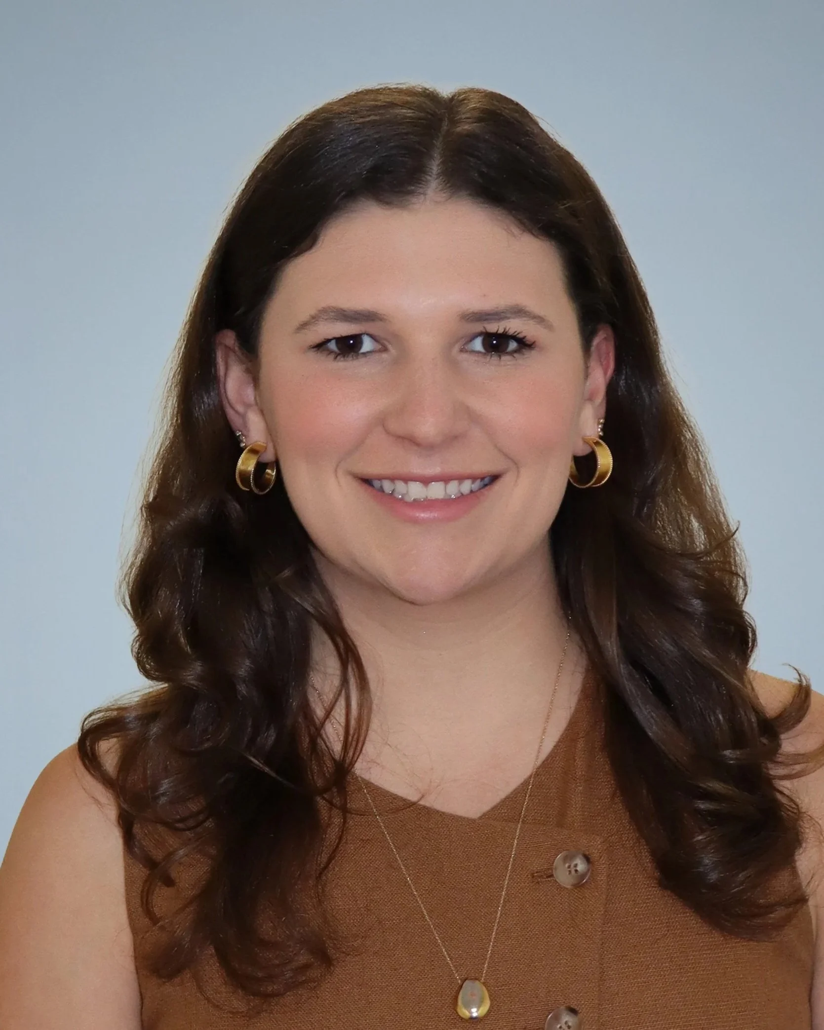 Close-up of a woman with long brown curly hair, wearing gold hoop earrings, a gold necklace with a pendant, and a sleeveless brown top, smiling against a plain light blue background.