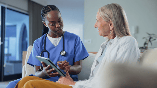 A nurse in blue scrubs showing information on a tablet to an elderly woman sitting on a hospital bed.