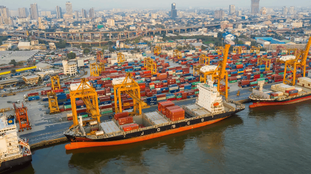 A busy port with cargo ships and yellow cranes loading and unloading containers, with a city skyline in the background.
