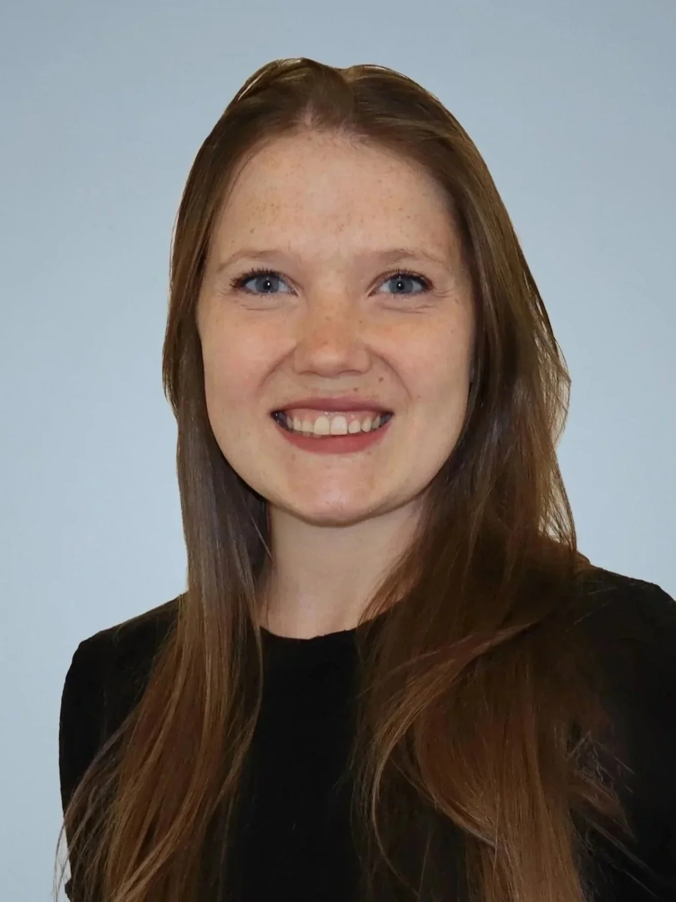 A young woman with long red hair, blue eyes, and freckles smiling against a light gray background.