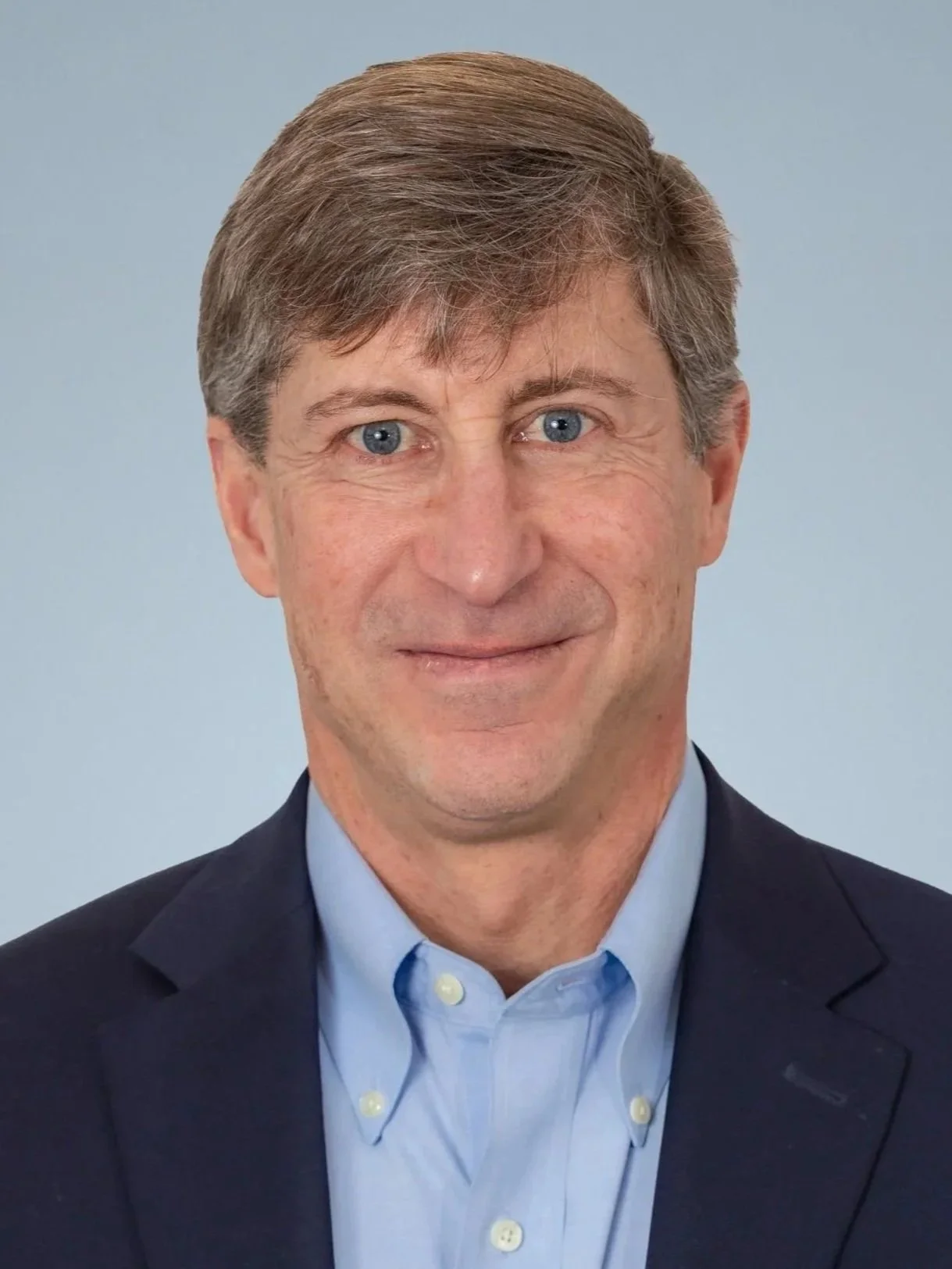A portrait of a middle-aged man with light brown hair and blue eyes, wearing a dark suit jacket over a light blue dress shirt, smiling against a plain light blue background.