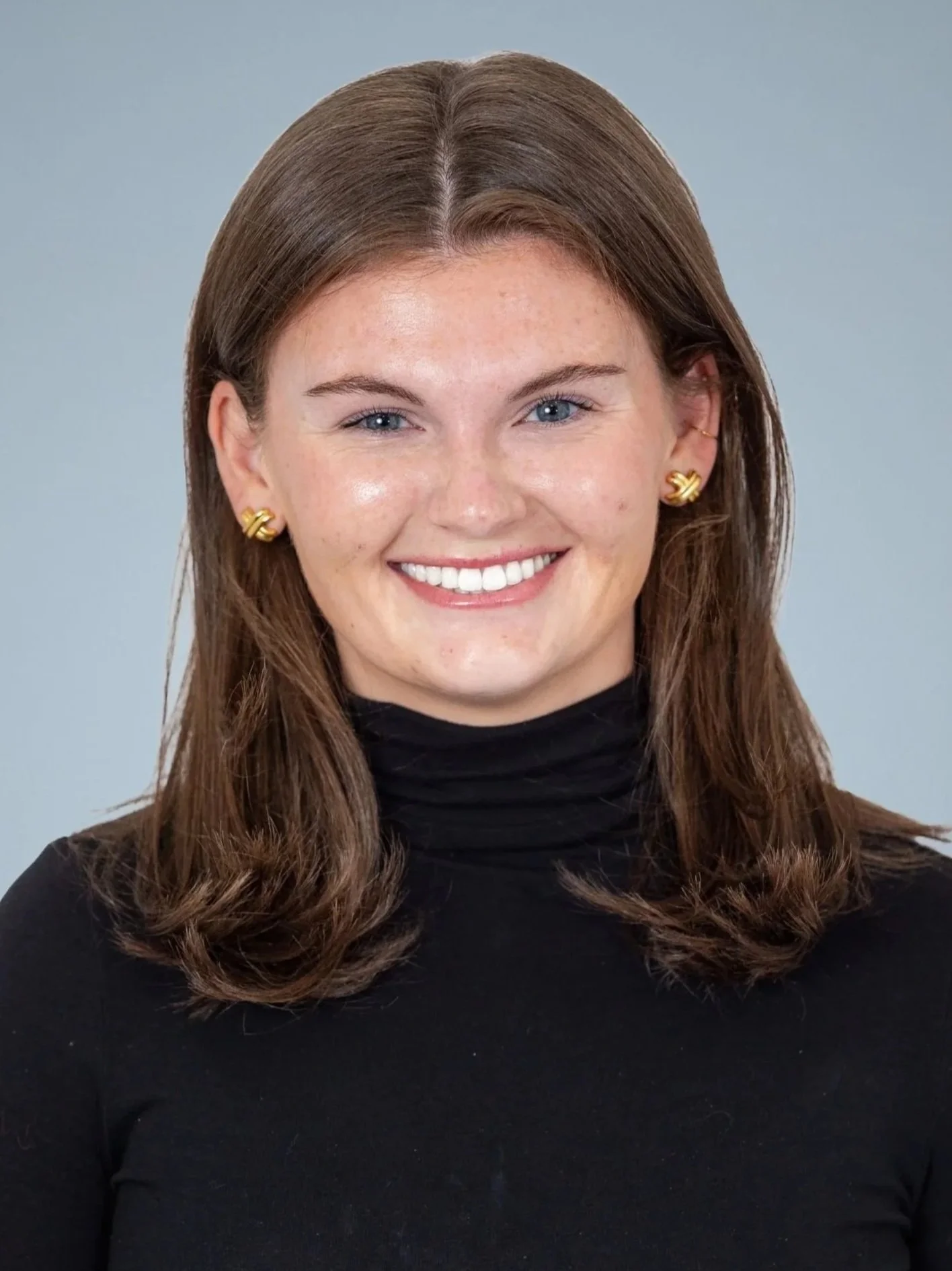 A woman with brown hair and blue eyes smiling, wearing gold earrings and a black turtleneck, against a light blue background.