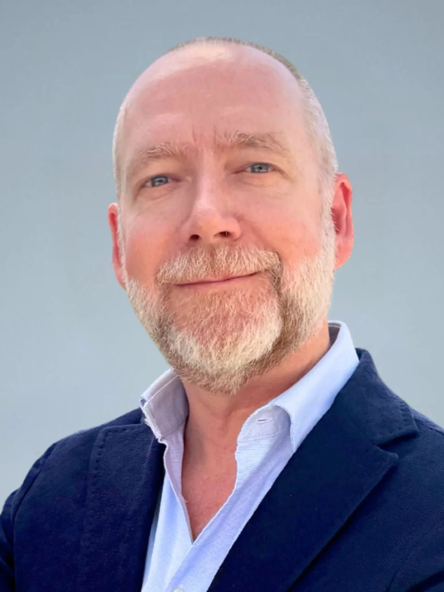 Close-up portrait of a middle-aged man with a beard, wearing a navy blue blazer and a light blue collared shirt, smiling against a plain gray background.