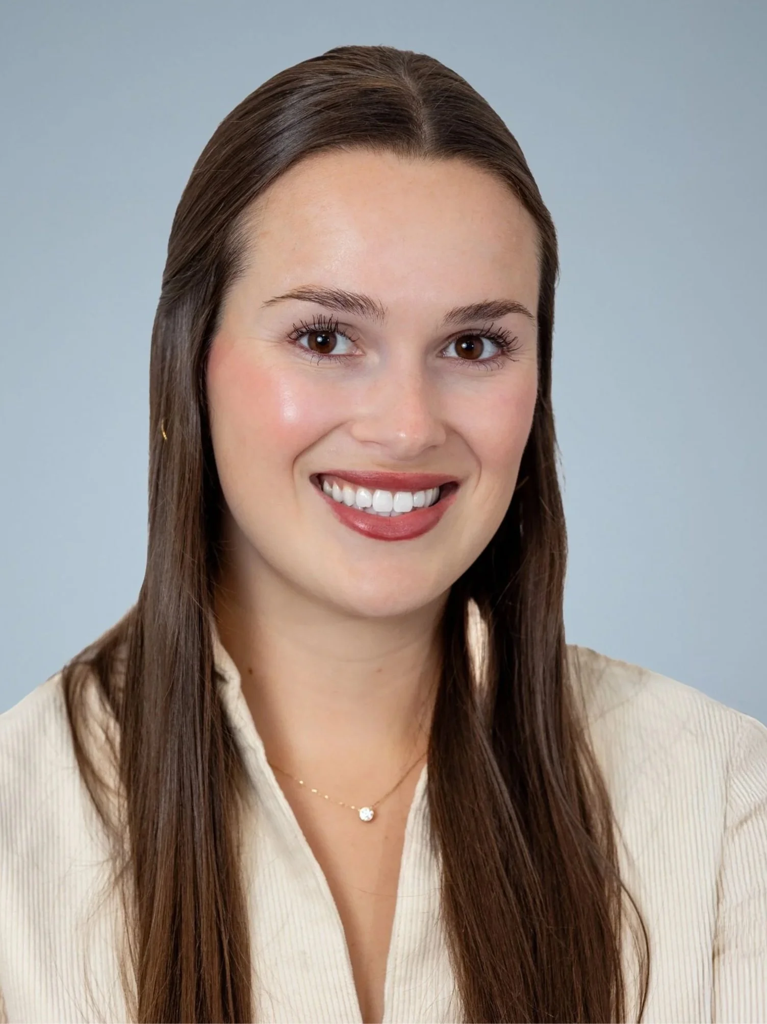 A woman smiling with brown hair, wearing a beige top and a delicate necklace, against a light blue background.