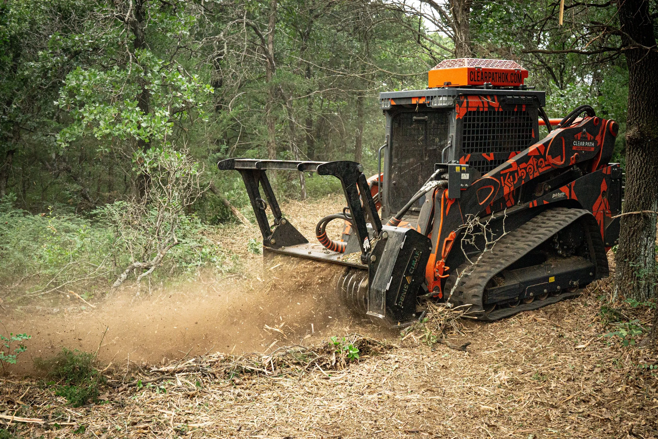 A track loader clearing dirt and debris from a wooded area surrounded by green trees and vegetation.