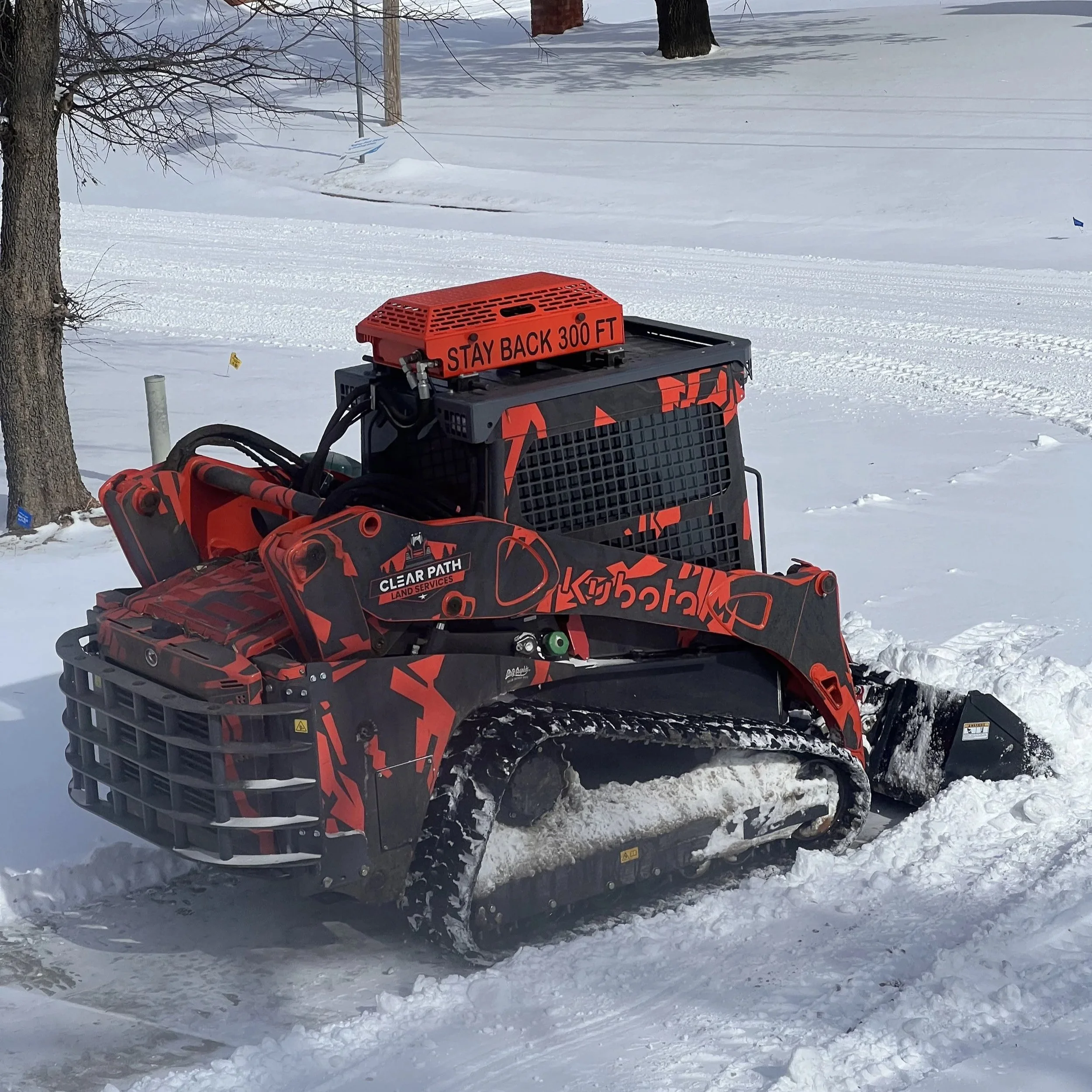 A snow-covered landscape with trees and electrical wires in the background, a red snowmobile in the foreground with a sign that reads 'Stay Back 300 Ft', and a small blue flag in the snow.