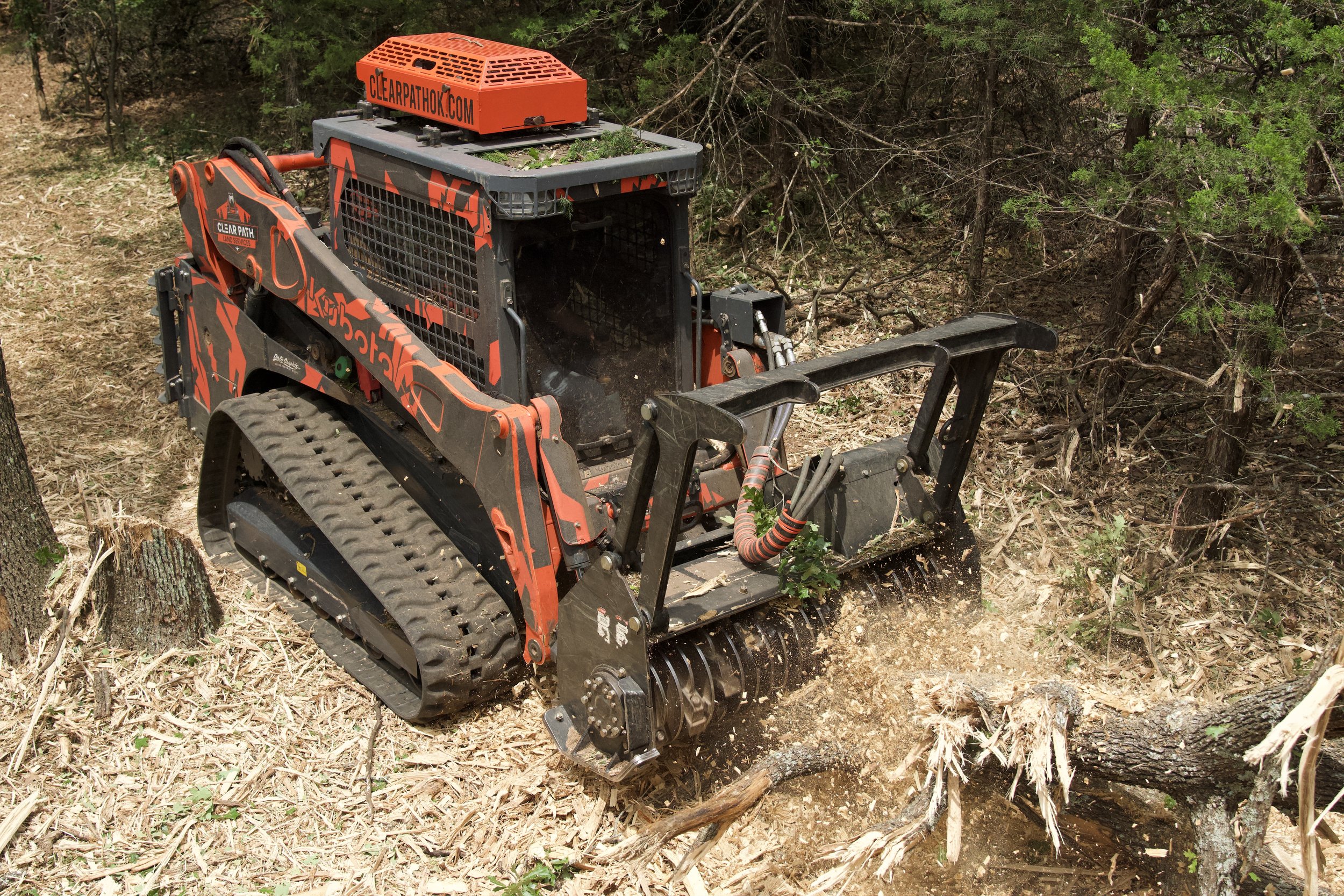 A tracked woodchipping machine with orange and black coloring working in a forested area, cutting through tree branches and wood debris.