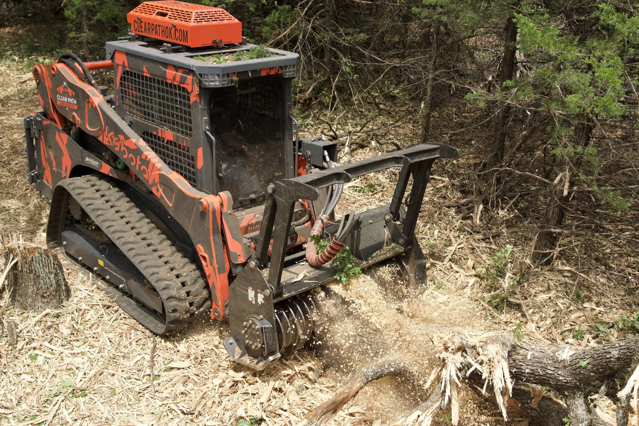Small robotic machine with tracks and a digging drum, working on clearing roots and debris in a wooded area.