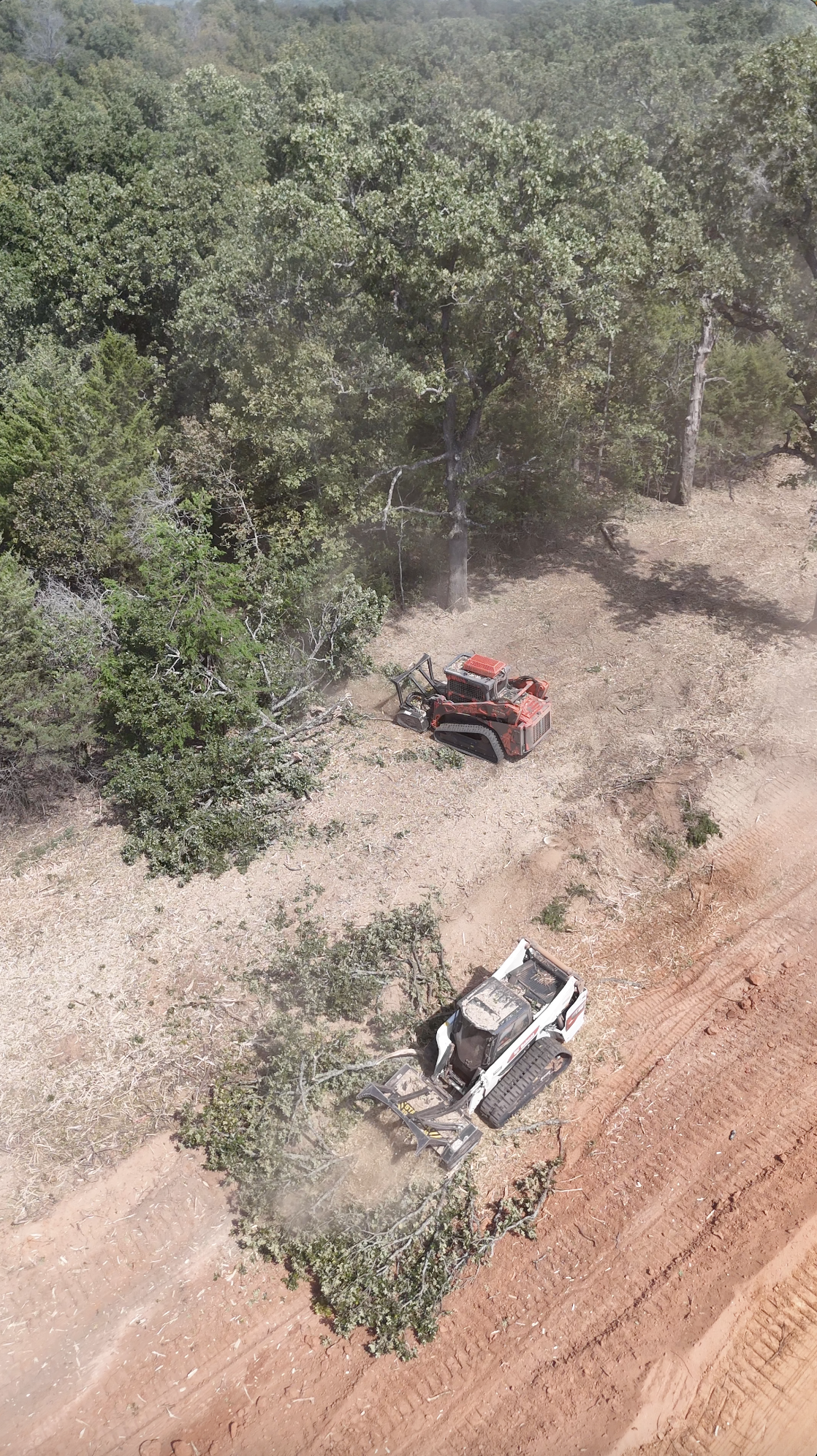 An aerial view of a construction site with two excavators, one red and one white, working among trees and dirt.