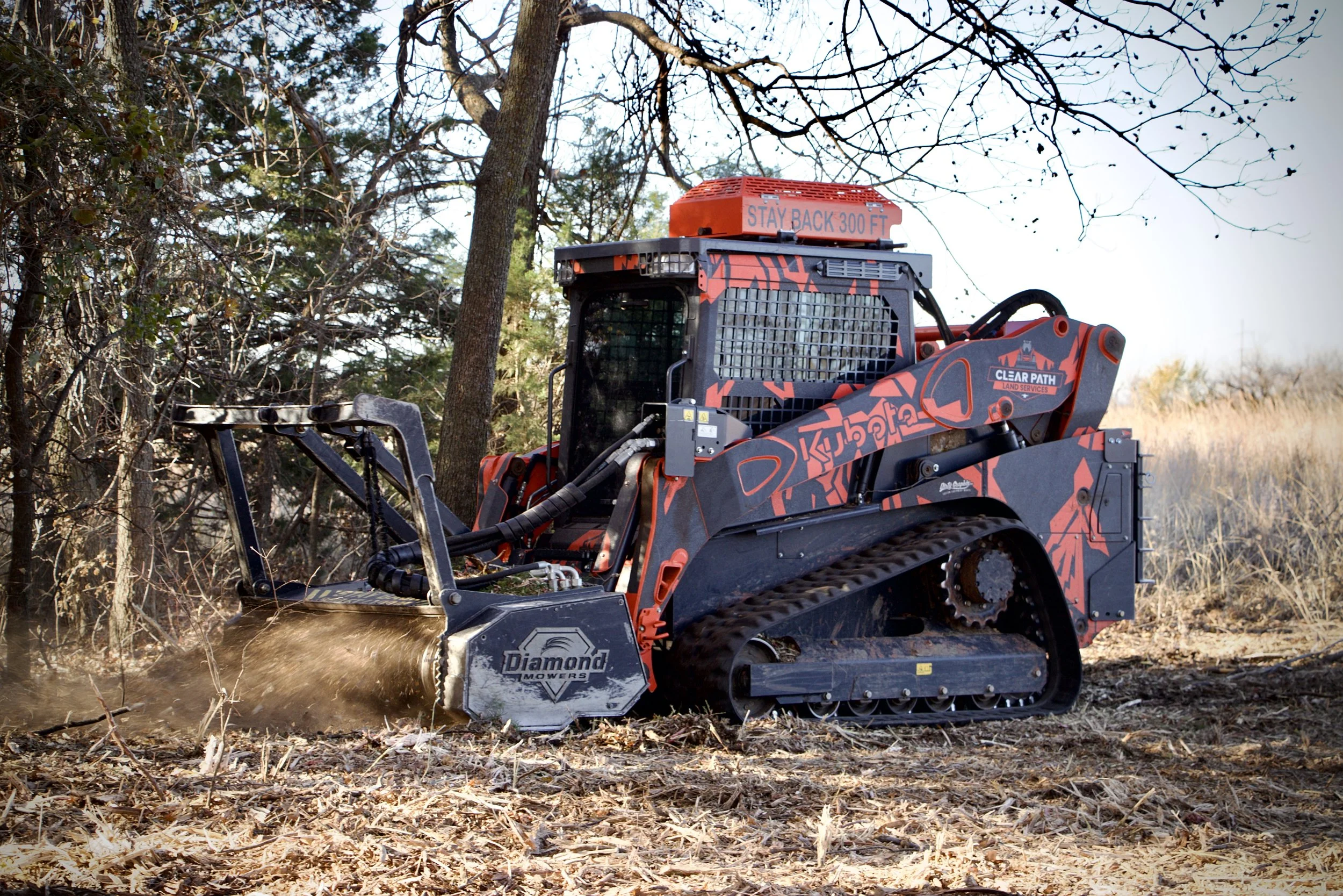 A skid steer loader working on a dirt clearing site, surrounded by trees and grass.
