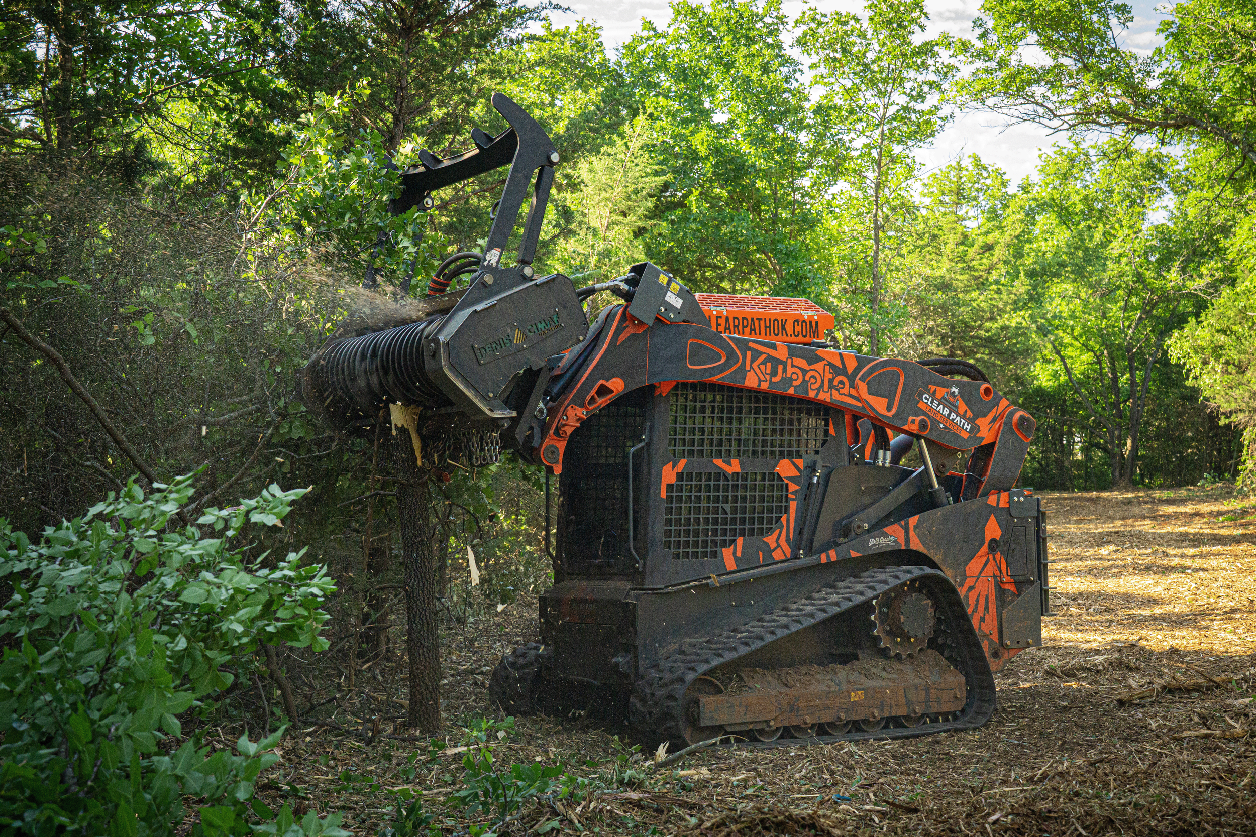 A compact excavator with an orange and black color scheme is being used to clear trees and brush in a wooded area. The machine has tracks and is equipped with a forestry attachment for trimming or removing branches.