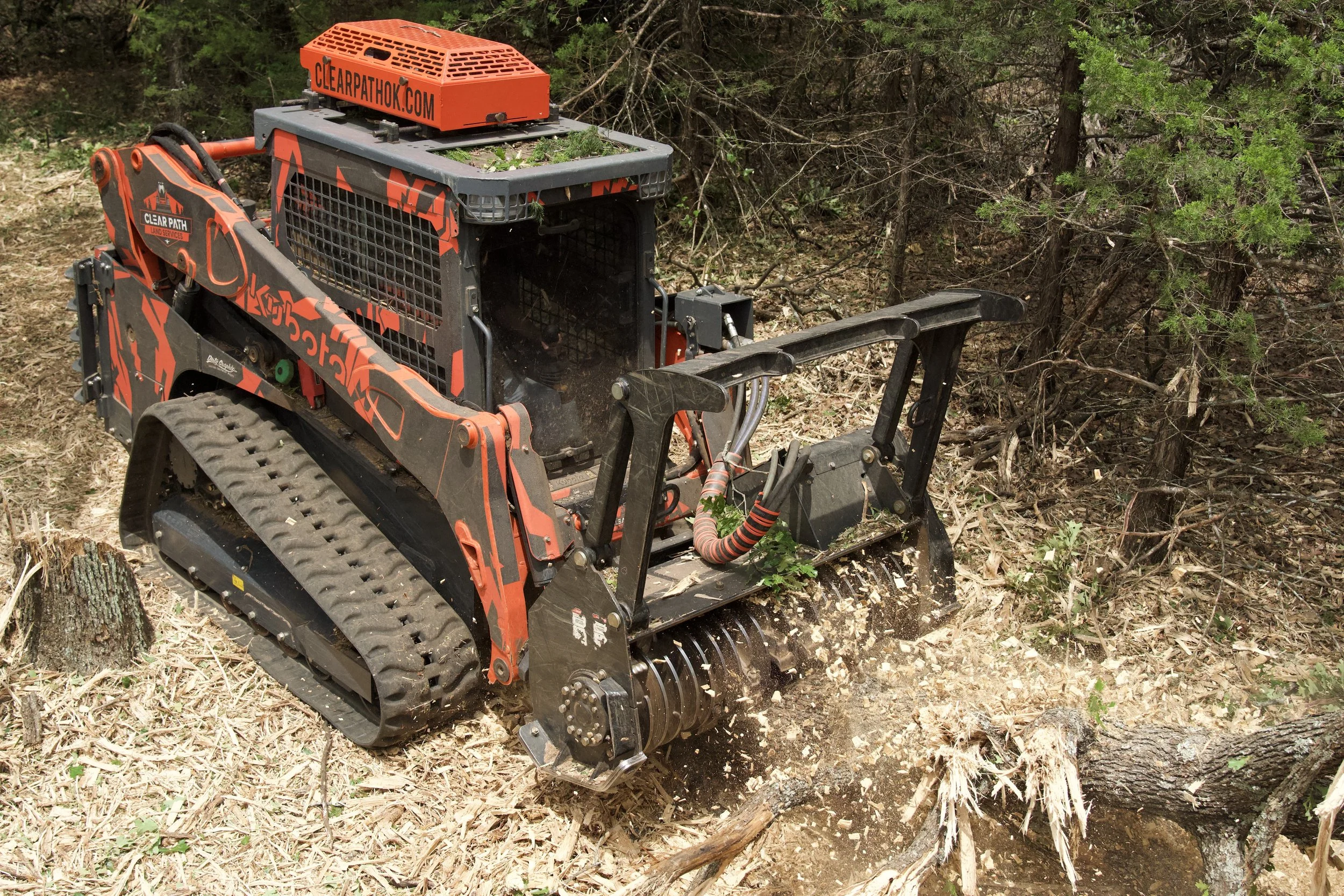 A tracked forestry mulcher in use in a forest clearing, grinding tree stumps and debris.