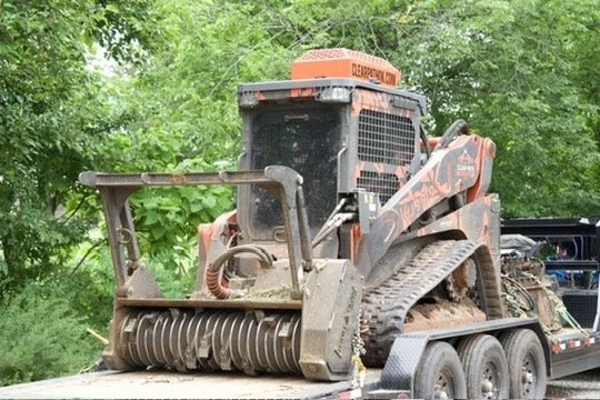 A small orange and black compact track loader on a trailer, surrounded by green trees.
