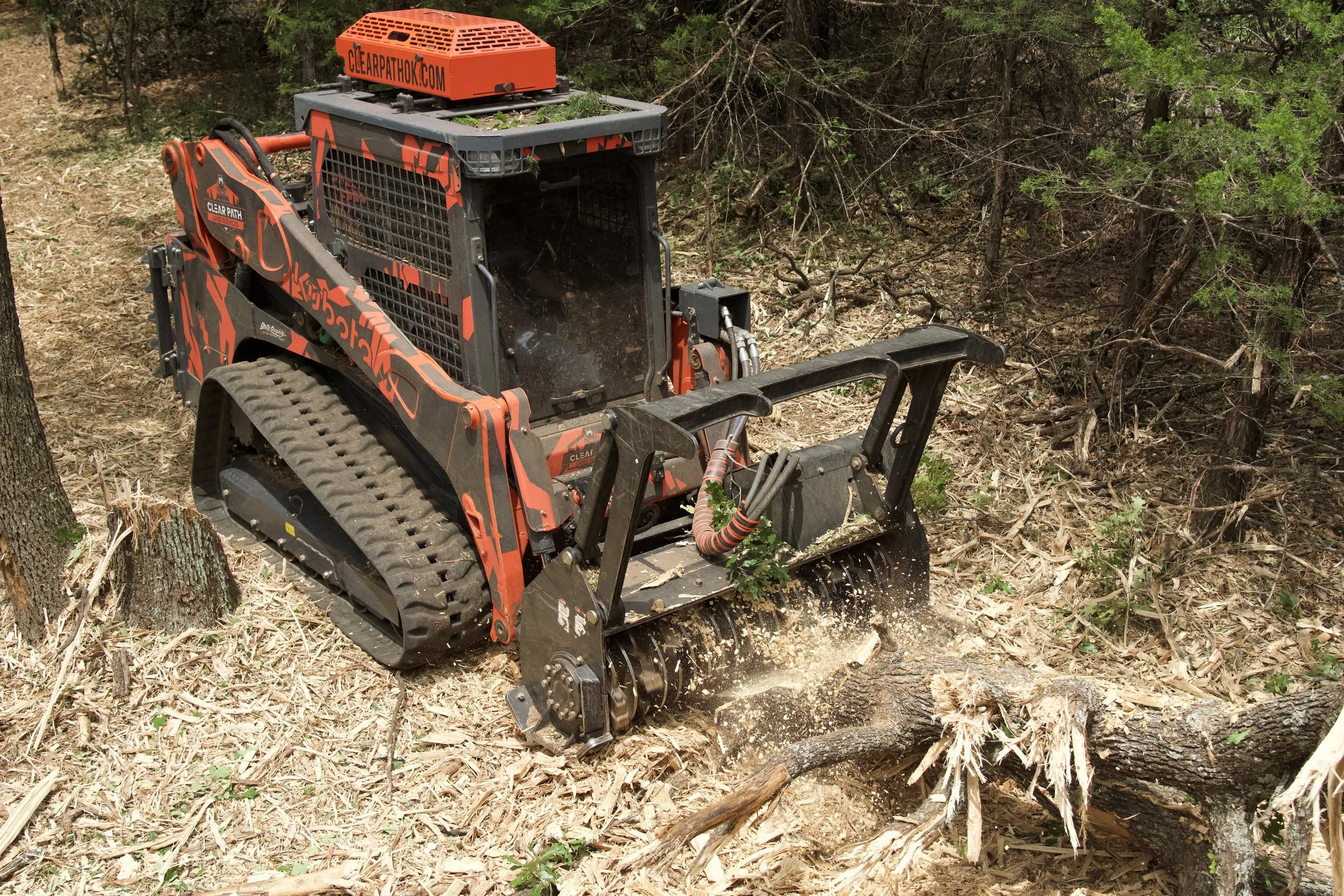 A tracked forestry mulcher attachment in orange and black on a small tractor operating in a wooded area, clearing debris and undergrowth.