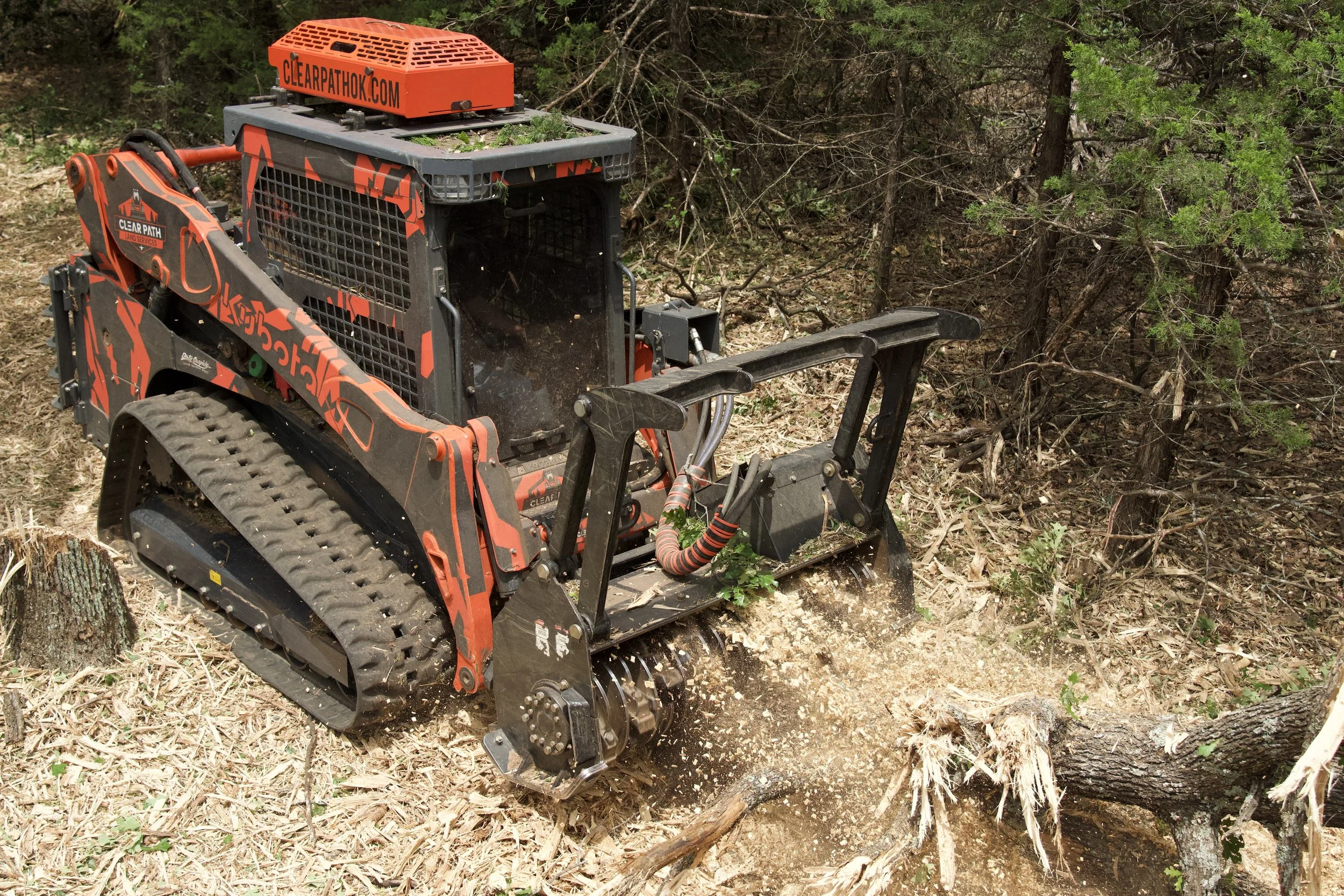 A compact woodchipper with tracks, orange and black coloring, and a metal grid enclosure, situated in a wooded area with trees and branches.