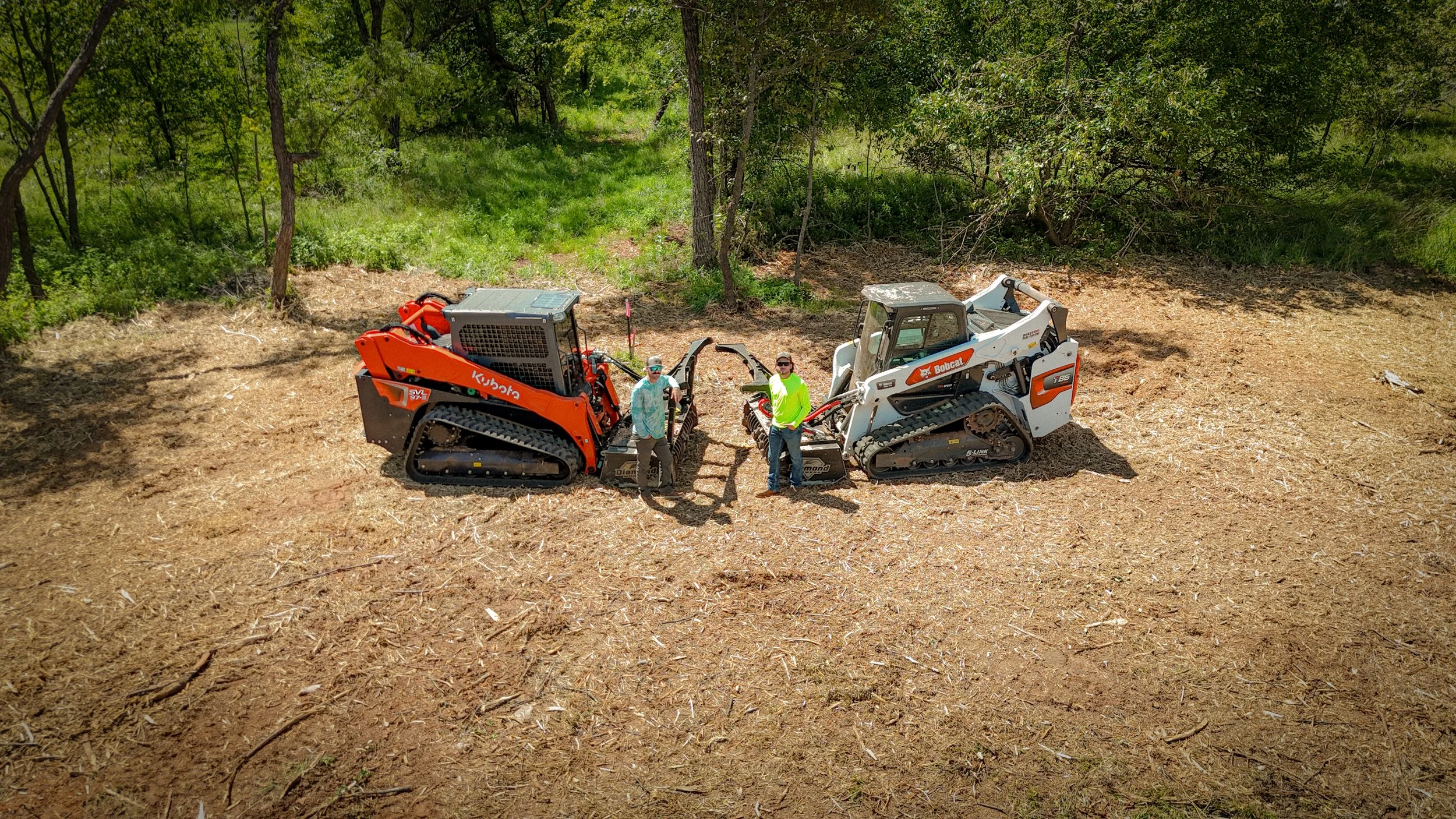 Two people standing between two tracked construction vehicles, one red and one white, on a cleared patch of ground surrounded by trees.